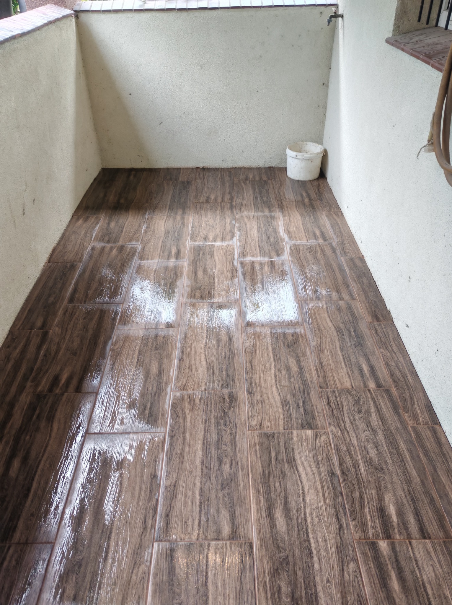 Błyszcząca, brązowa podłoga z płytek imitujących drewno na balkonie, świeżo po umyciu. Widoczny biały plastikowy kubeł i kran na ścianie.