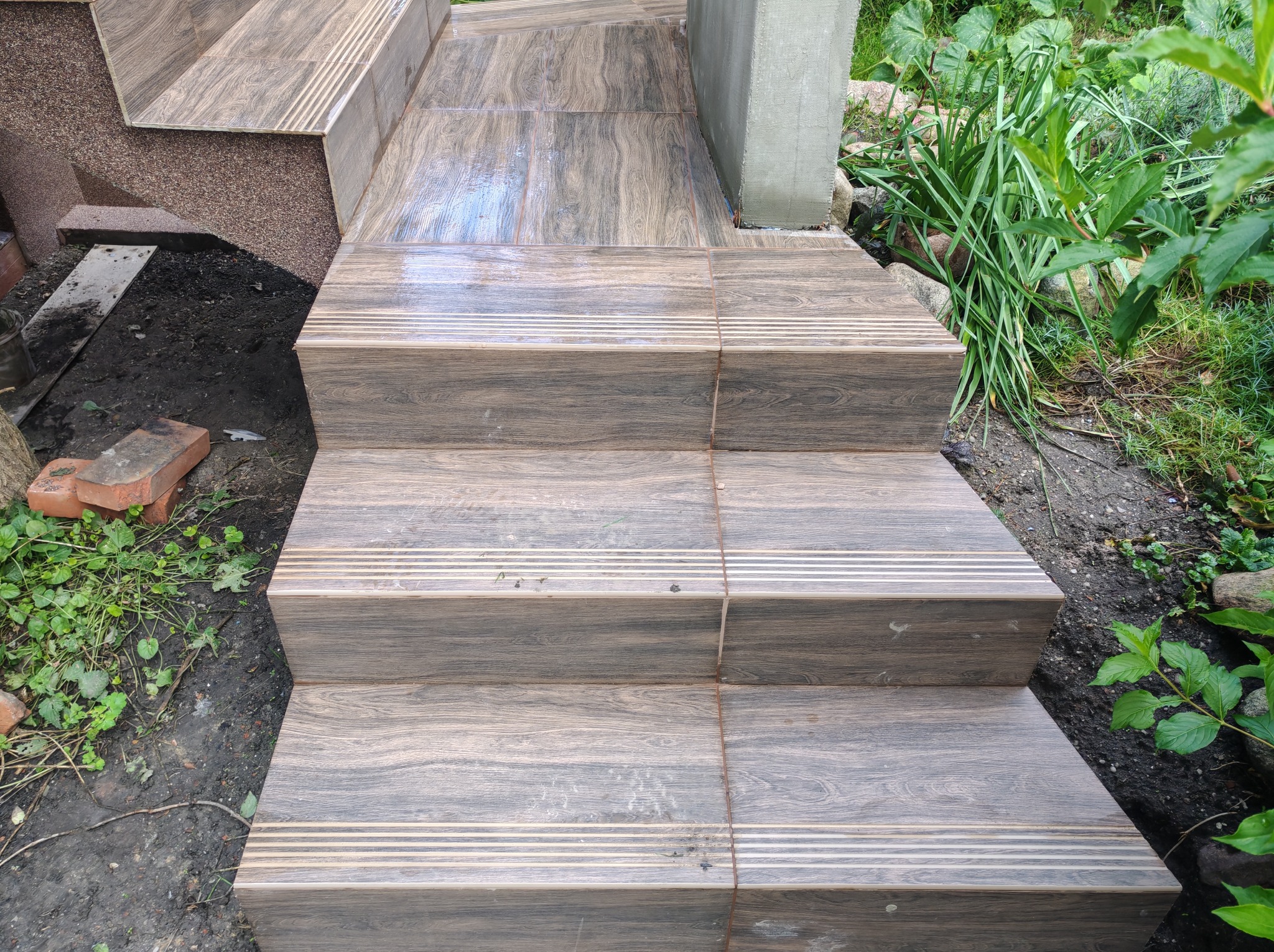 Schody zewnętrzne wyłożone brązowymi płytkami imitującymi drewno, z antypoślizgowymi paskami, prowadzące do ogrodu. Widoczne rośliny i elementy wykończeniowe.