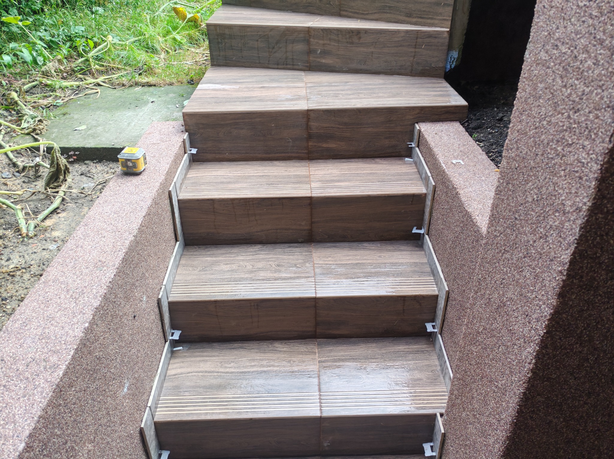 Schody zewnętrzne wyłożone brązowymi płytkami imitującymi drewno, z widocznymi elementami mocującymi. Ściany schodów wykończone tynkiem mozaikowym w odcieniach brązu.