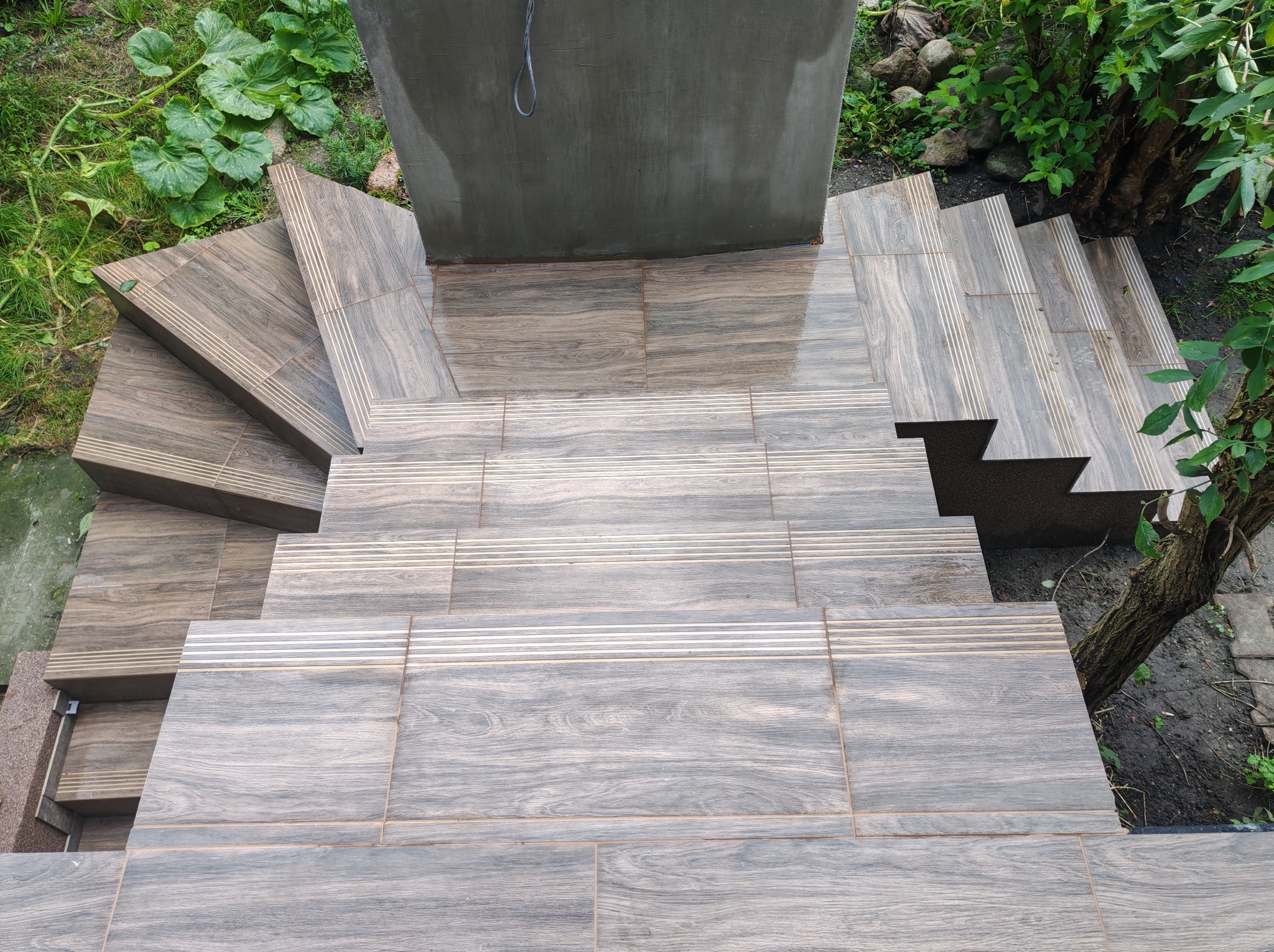 Wykończone płytkami schody zewnętrzne z efektem drewna, prowadzące w dół, otoczone zielenią. Widok z góry, ukazujący detale wzoru płytek i staranne ułożenie.