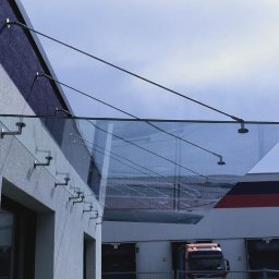 OwnGlass Oksana Boryslavska - Szklany daszek nad wejściem do budynku, mocowany na metalowych wspornikach. Widok z dołu, na tle błękitnego nieba i fragmentu budynku magazynowego.