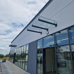 OwnGlass Oksana Boryslavska - Nowoczesny budynek z przeszkloną fasadą i szklanym zadaszeniem wspartym na czarnych, metalowych wspornikach. Widok z zewnątrz, na pierwszym planie chodnik z kostki.