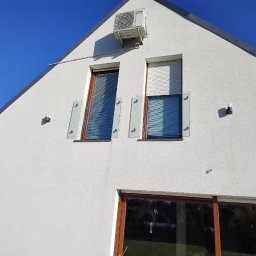 OwnGlass Oksana Boryslavska - Biały dom z dwoma oknami zabezpieczonymi szklanymi panelami. Na górze klimatyzator. Okna z drewnianymi ramami i roletami. Elewacja budynku w jasnym odcieniu.