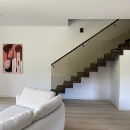 OwnGlass Oksana Boryslavska - Nowoczesne schody drewniane z balustradą ze szkła, widoczne z salonu. Minimalistyczne wnętrze z obrazem na ścianie i jasną podłogą.