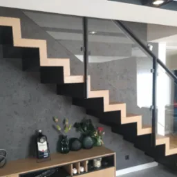 Nowoczesne schody wspornikowe z drewnianymi stopniami i szklaną balustradą, czarne elementy konstrukcyjne, dekoracyjny tynk na ścianie, oświetlenie punktowe.