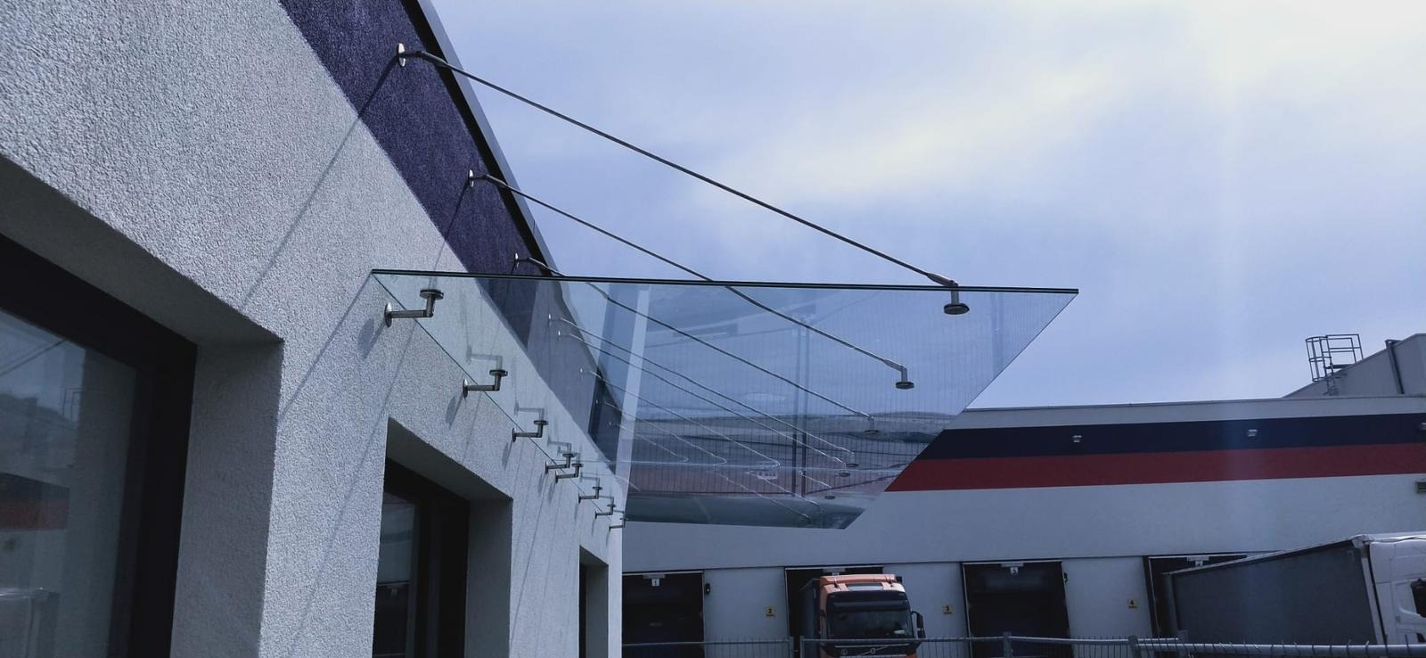 Szklany daszek nad wejściem do budynku, mocowany na metalowych wspornikach. Widok z dołu, na tle błękitnego nieba i fragmentu budynku magazynowego.
