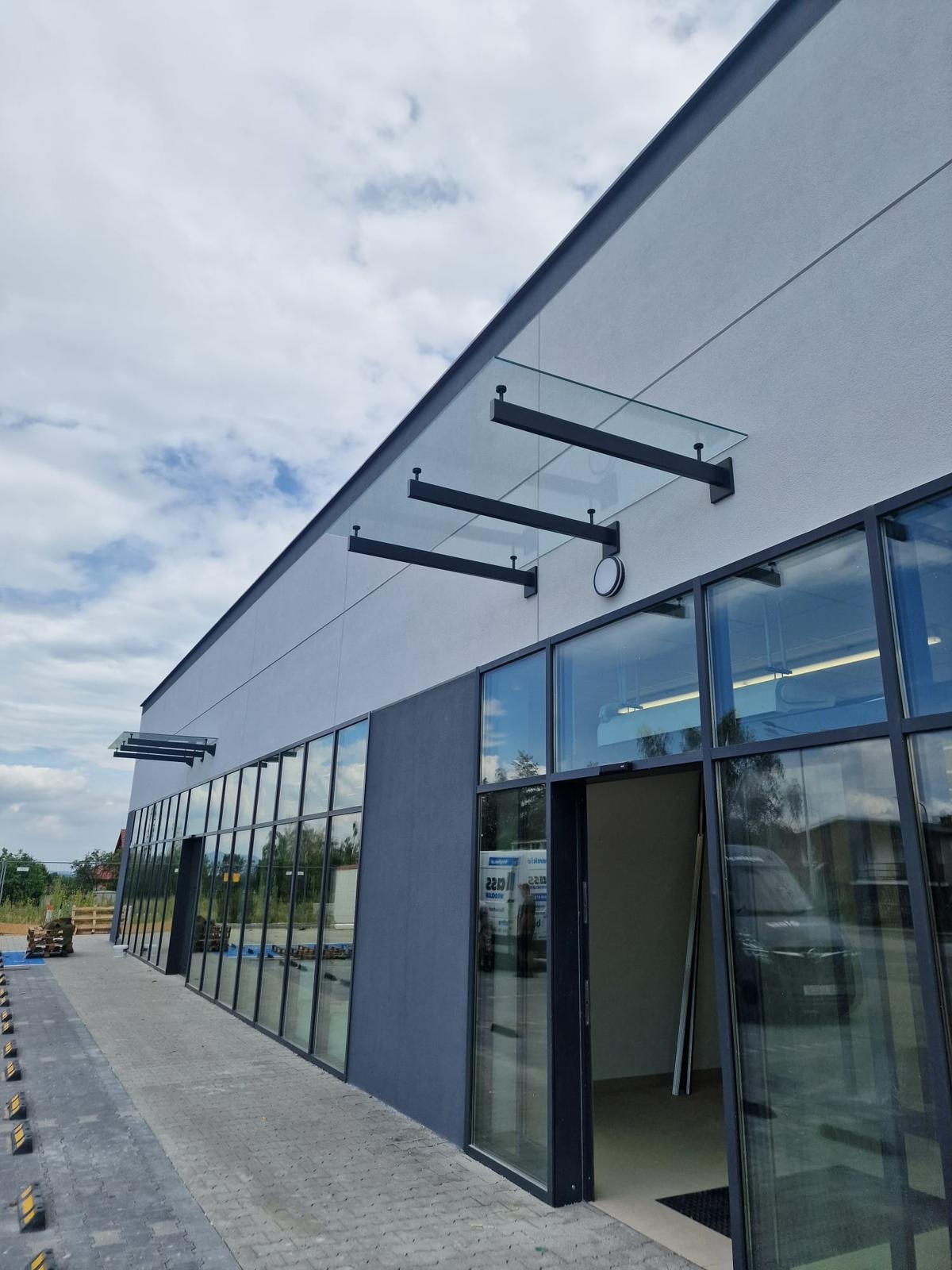 Nowoczesny budynek z przeszkloną fasadą i szklanym zadaszeniem wspartym na czarnych, metalowych wspornikach. Widok z zewnątrz, na pierwszym planie chodnik z kostki.