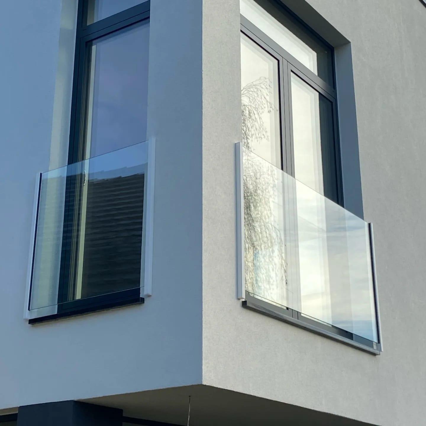 Nowoczesna, szklana balustrada balkonowa z grafitowymi ramami, zamontowana na elewacji budynku o jasnoszarym tynku strukturalnym. Widok z dołu, podkreślający minimalistyczny design.