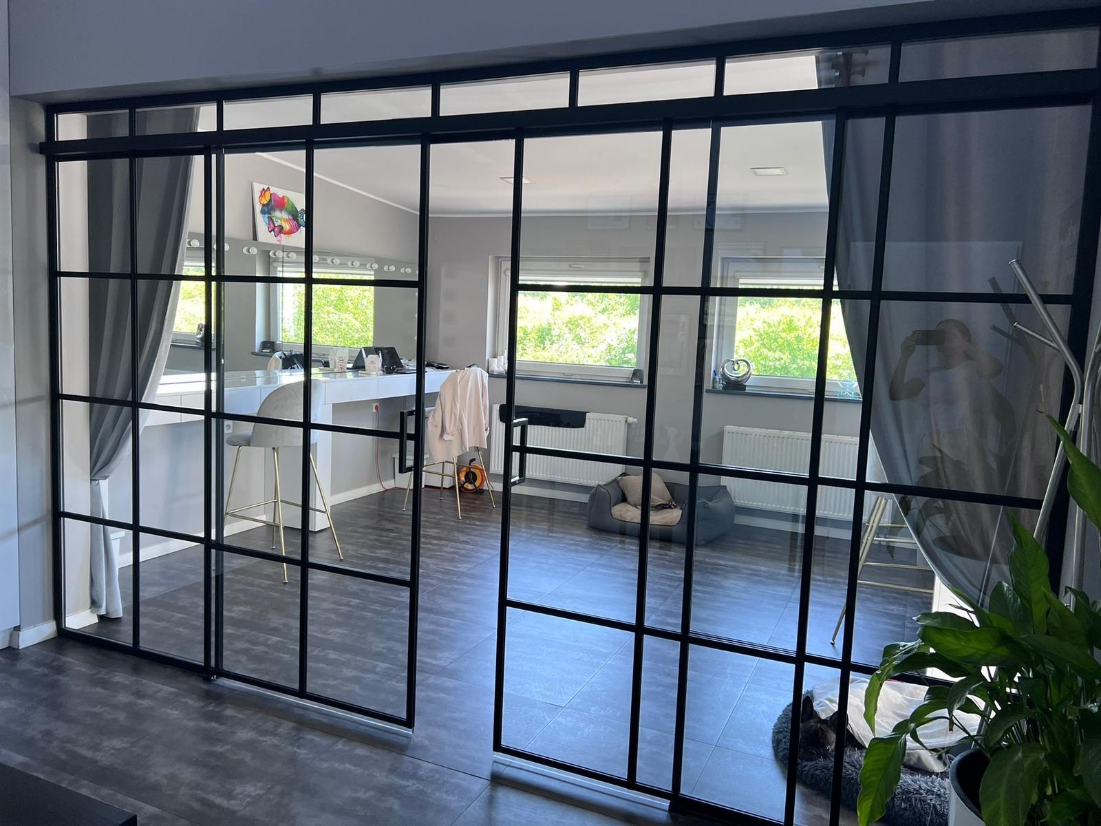 Przeszklona ściana działowa z czarnymi szprosami oddziela pokoje. Widoczna toaletka z lustrem i krzesłem. W tle okno z widokiem na zieleń. Minimalistyczny design.