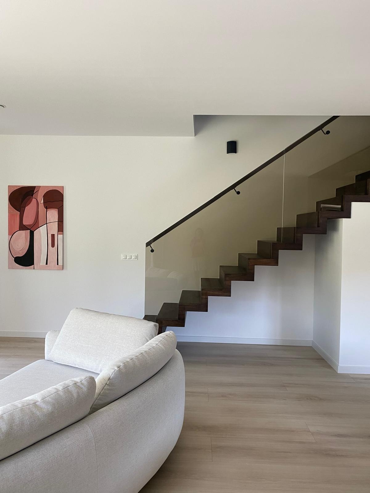 Nowoczesne schody drewniane z balustradą ze szkła, widoczne z salonu. Minimalistyczne wnętrze z obrazem na ścianie i jasną podłogą.