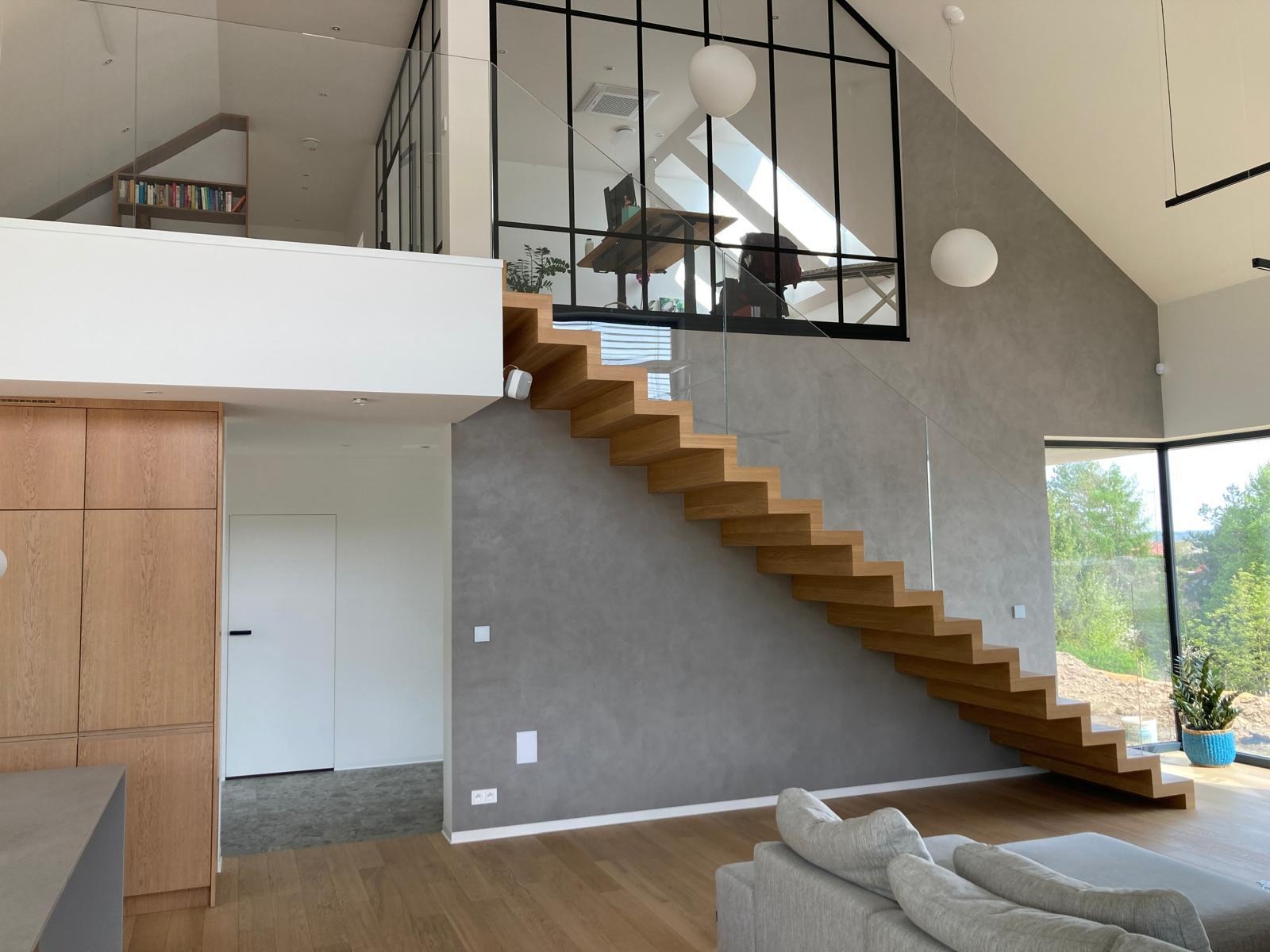 Nowoczesne schody drewniane z balustradą szklaną w przestronnym wnętrzu domu w Jeszkowicach. Widoczna antresola z przeszkloną ścianą.