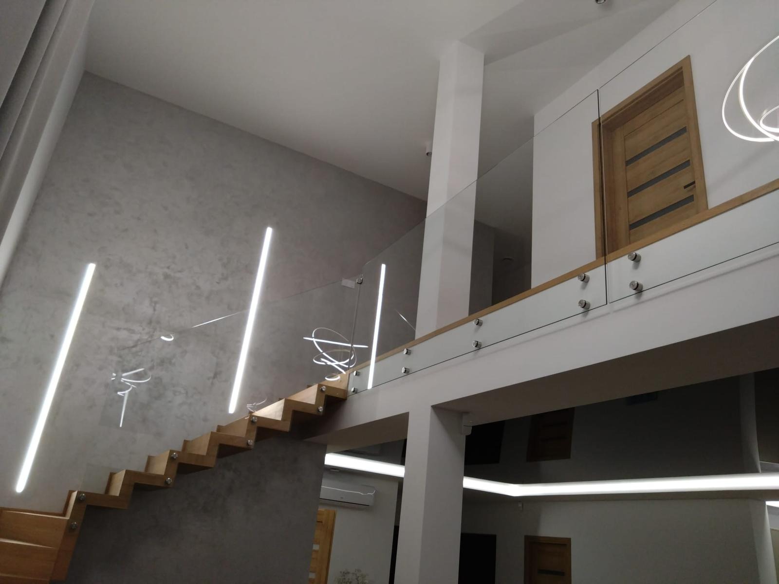 Nowoczesne wnętrze z drewnianymi schodami i szklaną balustradą mocowaną punktowo. Minimalistyczny design, oświetlenie LED. Widok na antresolę i drewniane drzwi.
