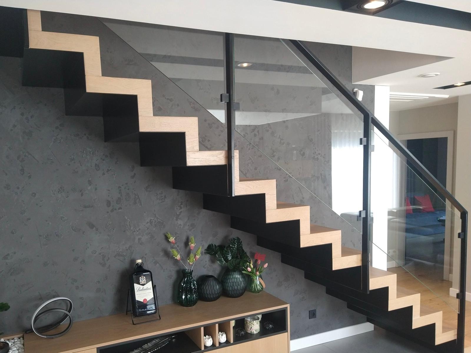 Nowoczesne schody wspornikowe z drewnianymi stopniami i szklaną balustradą, czarne elementy konstrukcyjne, dekoracyjny tynk na ścianie, oświetlenie punktowe.