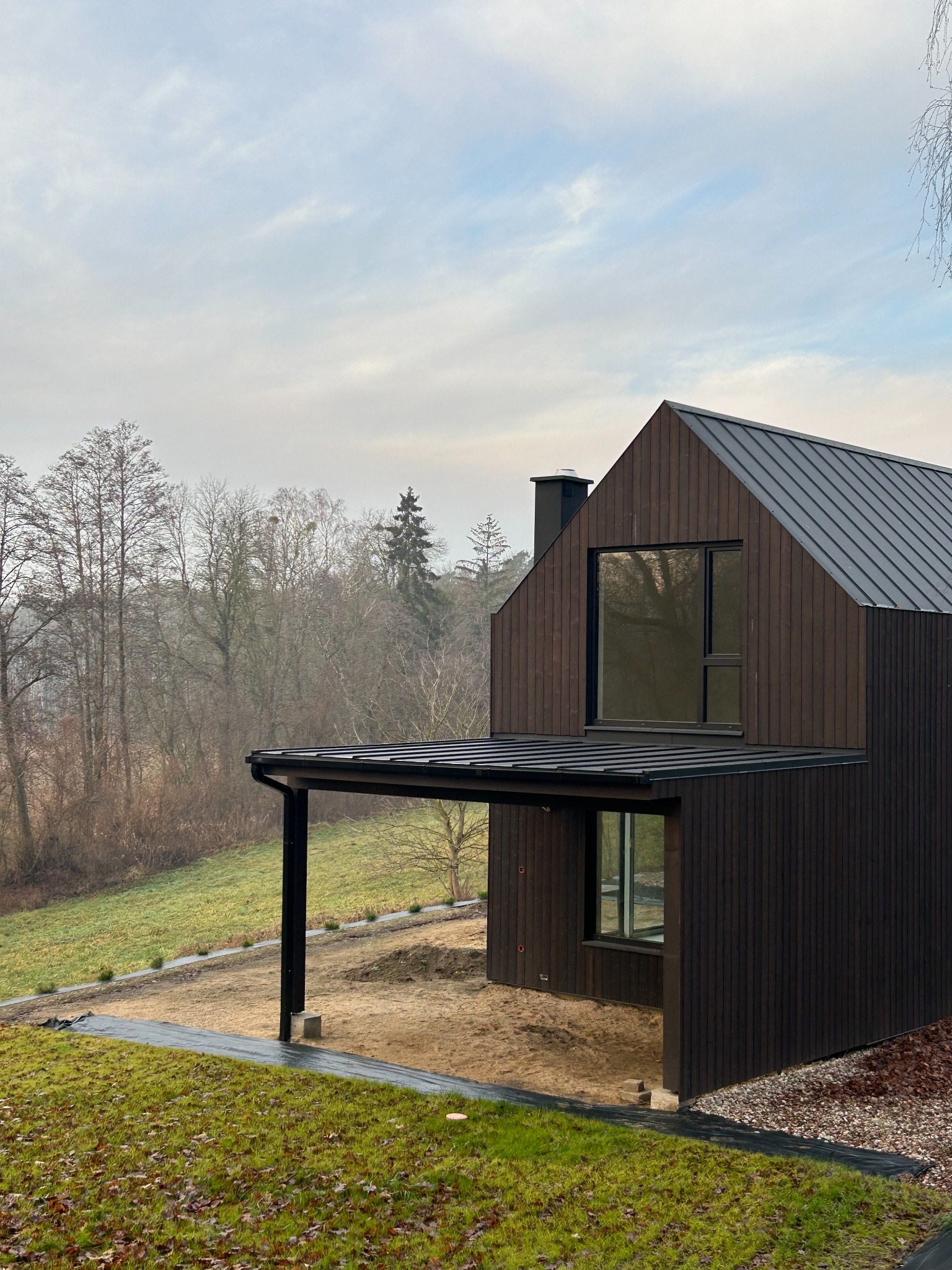 Nowoczesny dom z ciemnego drewna z płaskim dachem i zadaszonym tarasem. Minimalistyczny design, otoczenie naturalne, trawa i drzewa. Widok z dołu.