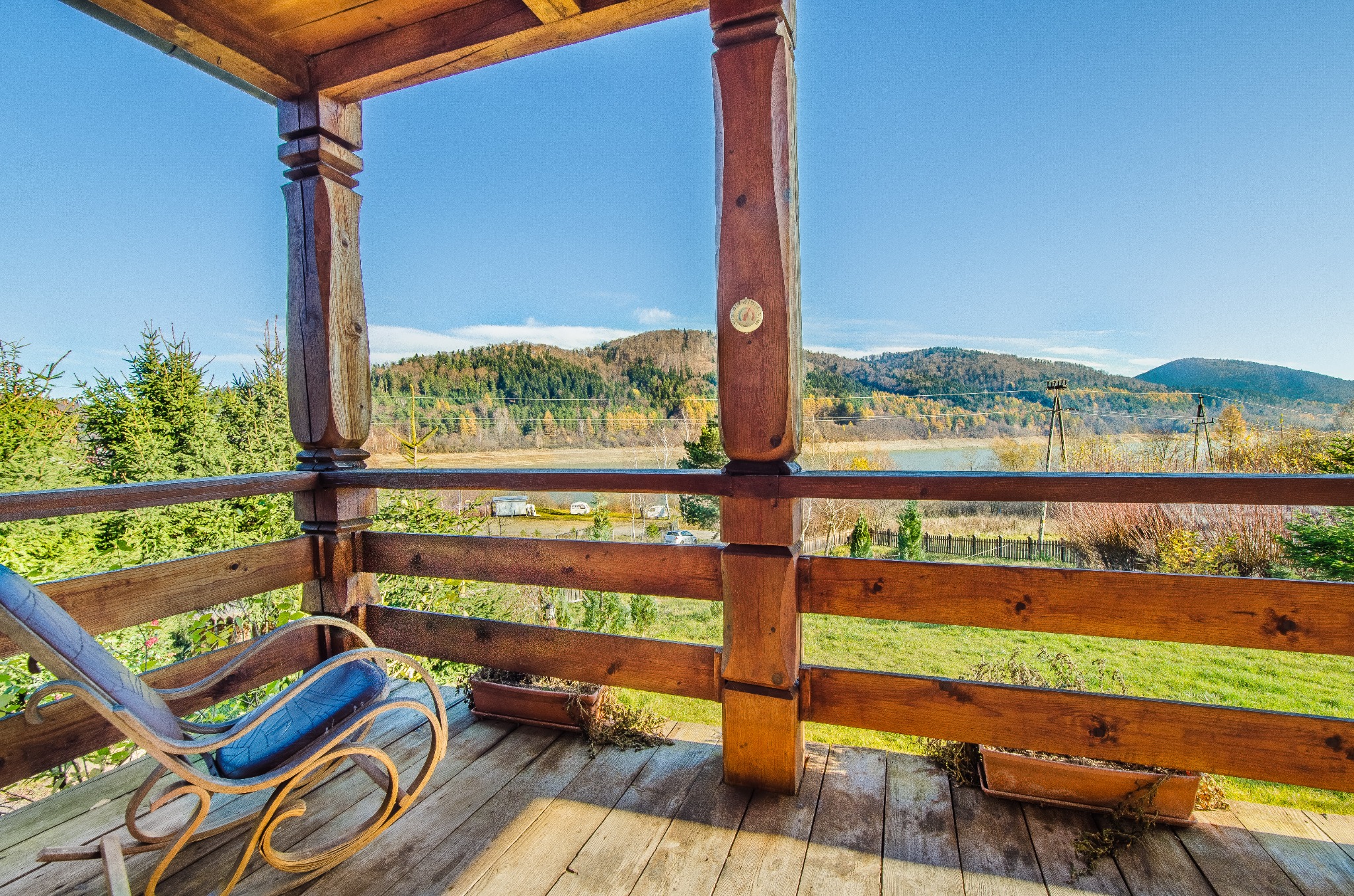 Rustykalny balkon z drewnianą balustradą i widokiem na jezioro i góry w słoneczny dzień. Na podłodze drewniany fotel bujany.