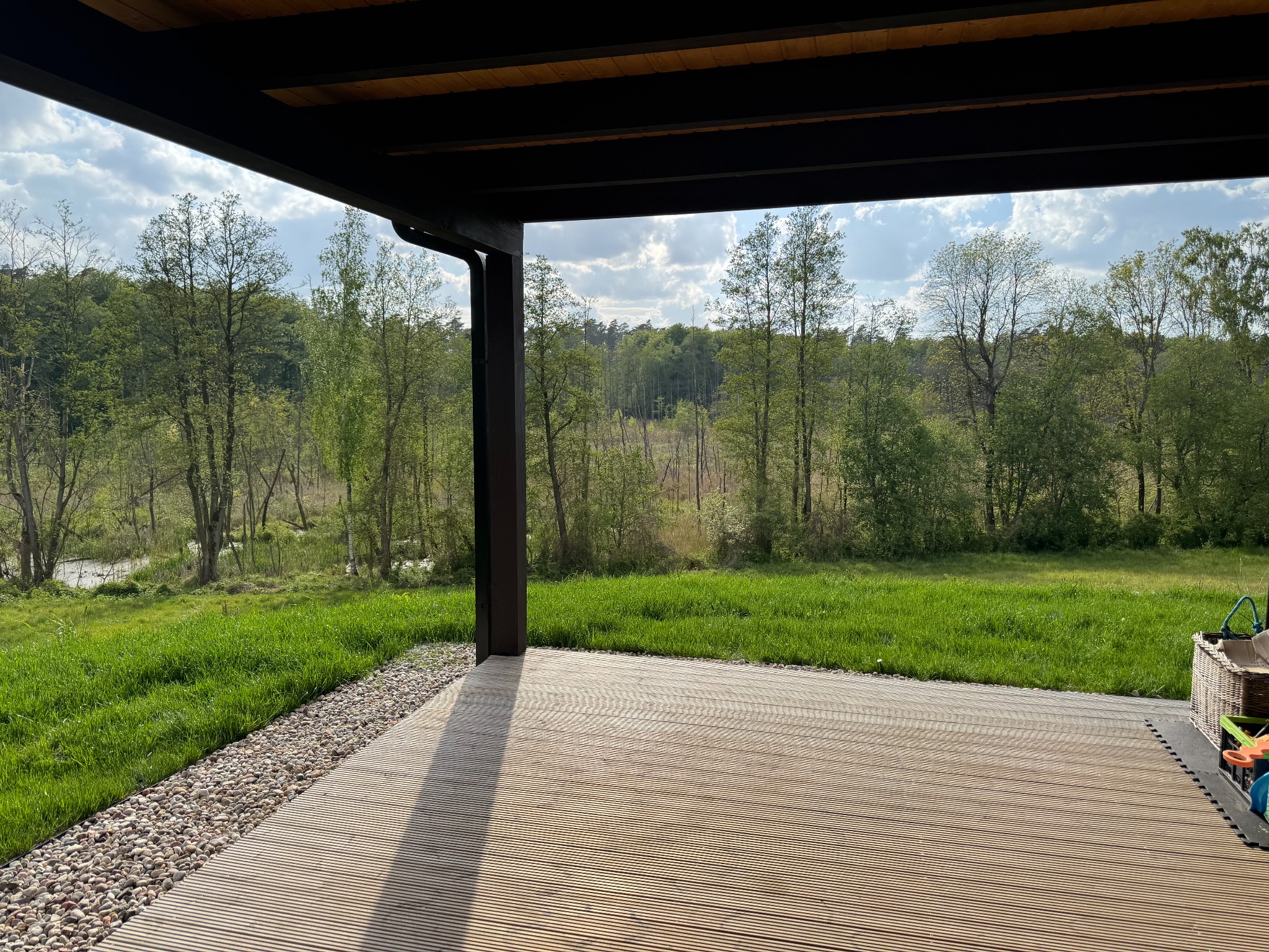 Zadaszony taras z drewnianą podłogą i widokiem na zielony krajobraz z drzewami i łąką w słoneczny dzień. Kamienista rabata oddziela taras od trawnika.