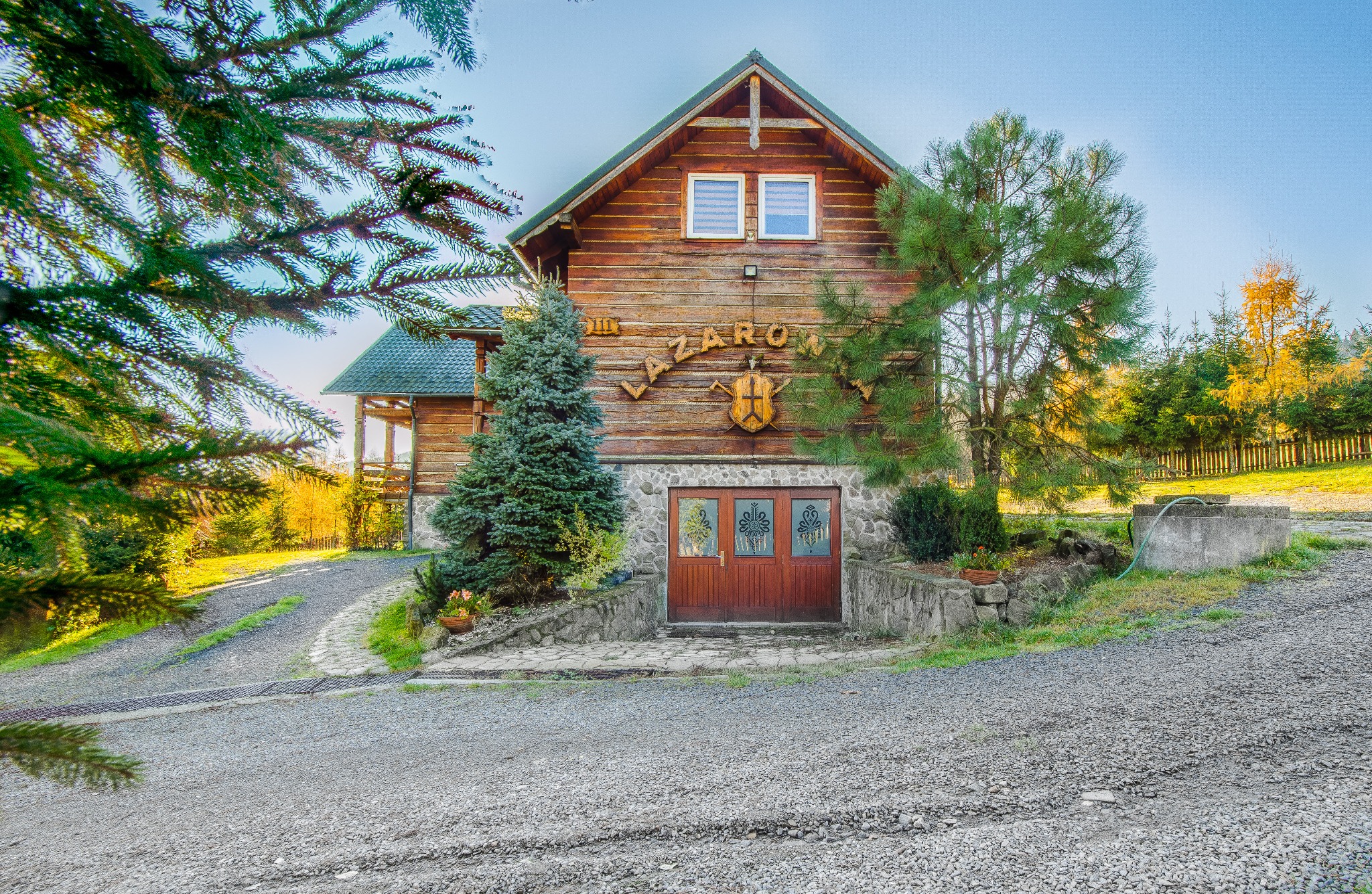 Drewniany dom z napisem 'Łazarowy' i ozdobnym herbem, otoczony zielenią, prezentuje się rustykalnie i przytulnie. Kamienna podmurówka i ścieżka dodają uroku.