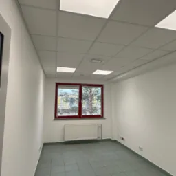 Puste, jasne pomieszczenie z szarą podłogą z płytek, białymi ścianami, oknem z czerwoną ramą i grzejnikiem. Sufit podwieszany z kwadratowymi panelami.