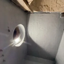 Szary drenaż studzienki z oświetleniem wewnątrz, widoczne ściany i sufit o nierównej fakturze. Kąt ujęcia z dołu, podkreślający głębię konstrukcji.