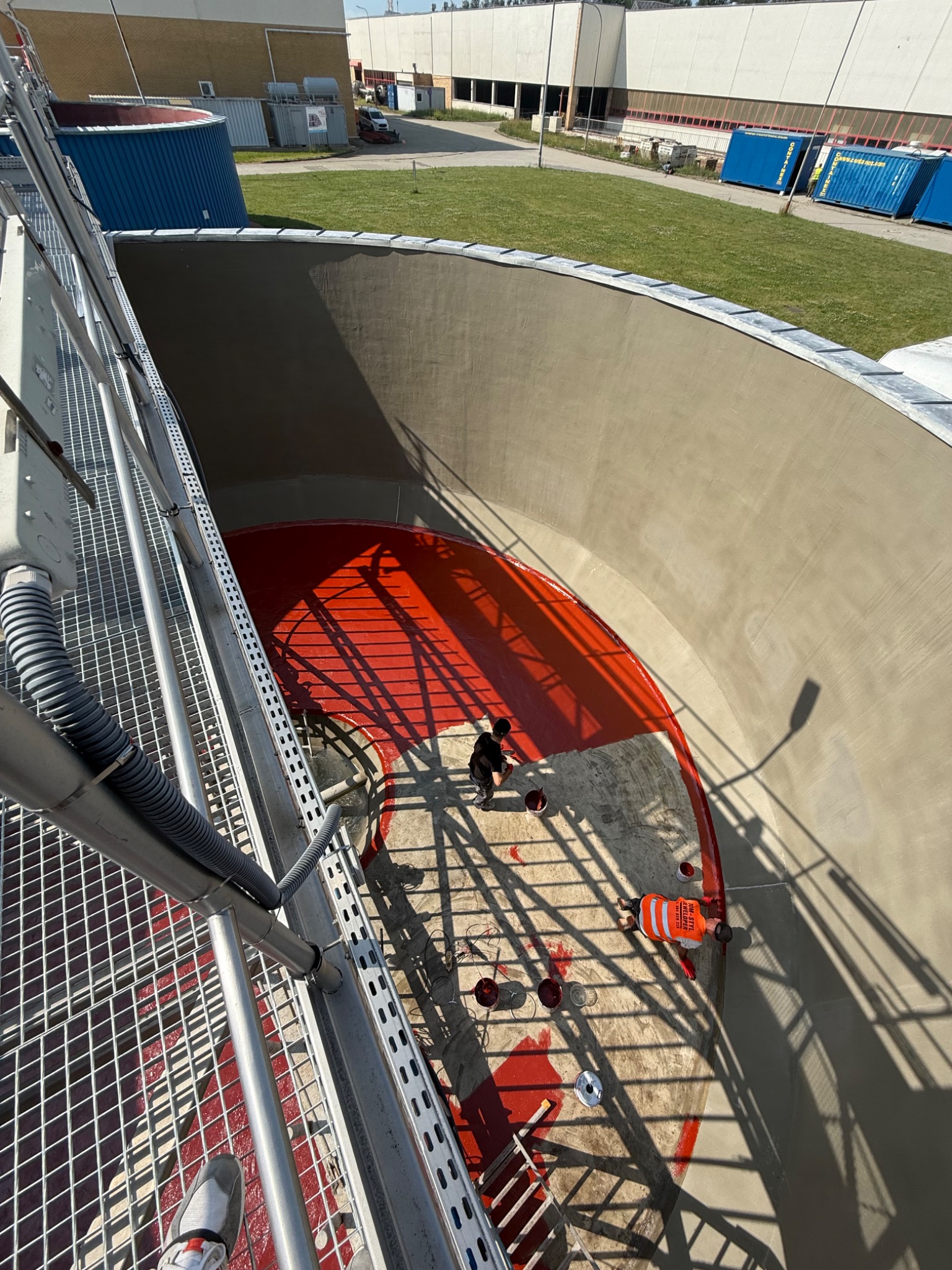 Malowanie wnętrza betonowego zbiornika na czerwono z metalowej platformy. Widoczni pracownicy w odblaskowych kamizelkach i puszki z farbą.