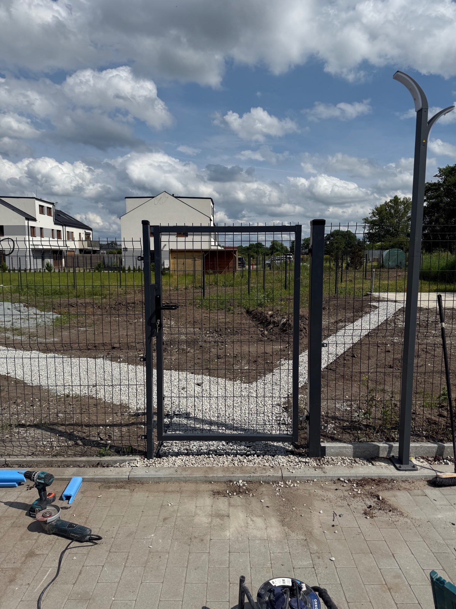 Montaż ogrodzenia panelowego z furtką na posesji. Widoczne narzędzia i ścieżka z kamienia. Nowoczesne budynki w tle, pochmurne niebo.