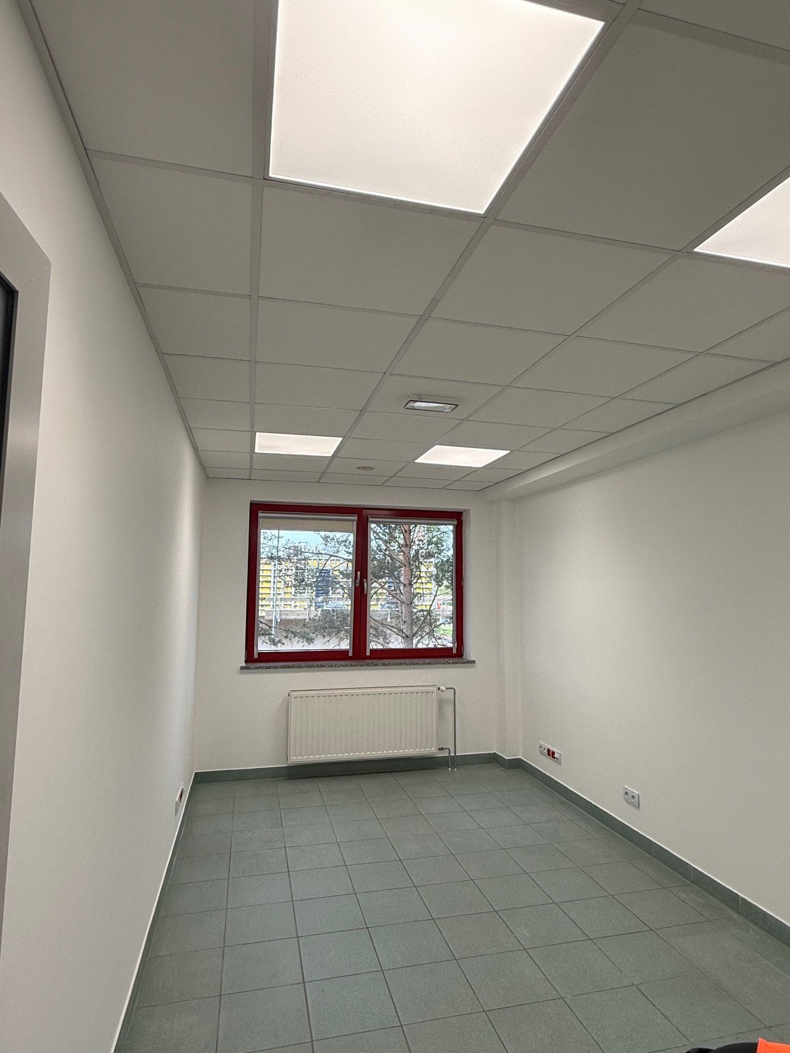 Puste, jasne pomieszczenie z szarą podłogą z płytek, białymi ścianami, oknem z czerwoną ramą i grzejnikiem. Sufit podwieszany z kwadratowymi panelami.