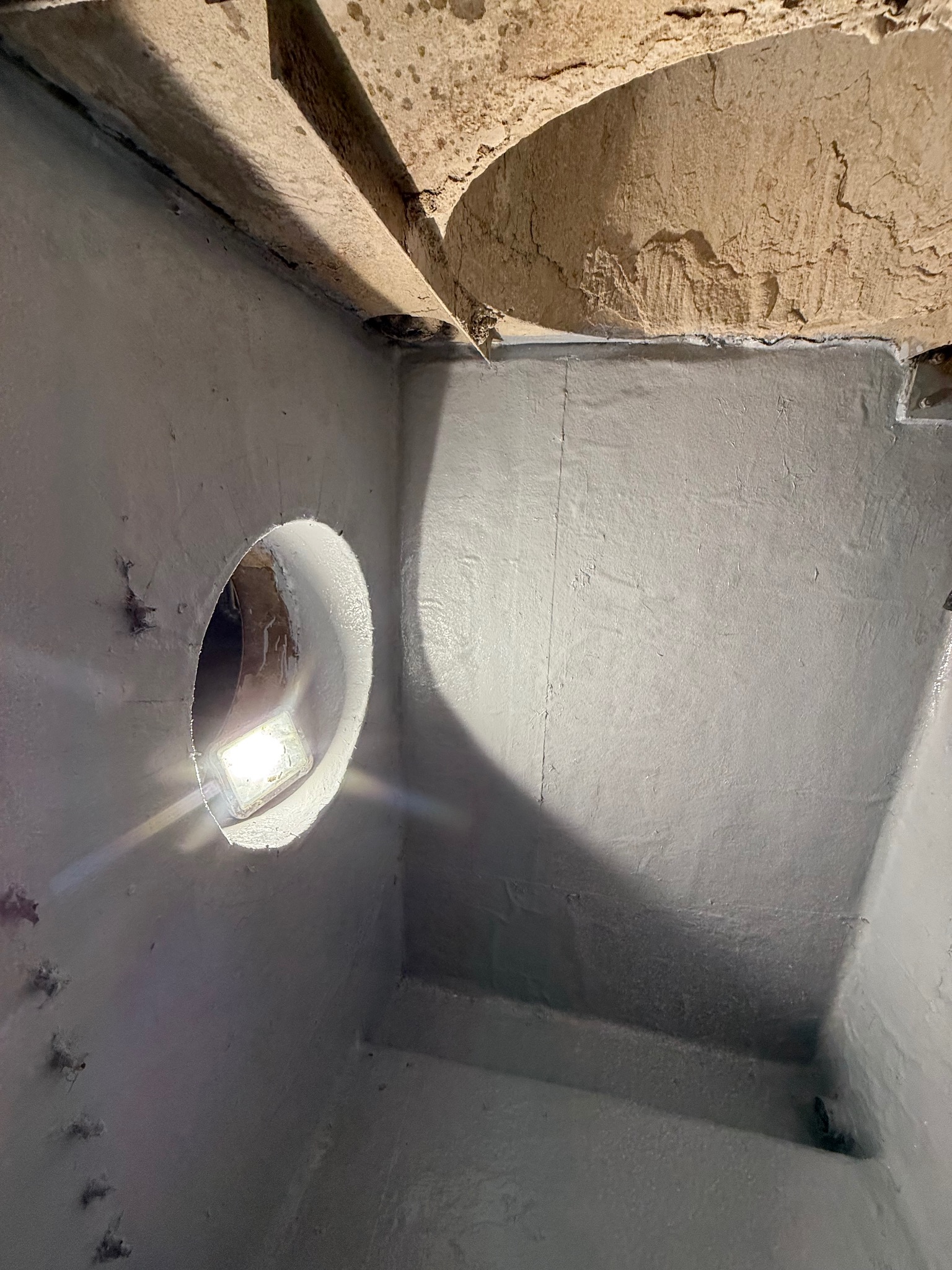 Szary drenaż studzienki z oświetleniem wewnątrz, widoczne ściany i sufit o nierównej fakturze. Kąt ujęcia z dołu, podkreślający głębię konstrukcji.