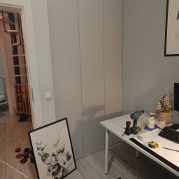 Okperfectlymade - Jasnoszara szafa wnękowa z trzema frontami, widoczna w trakcie montażu z narzędziami na biurku i dekoracją. W tle schody spiralne.