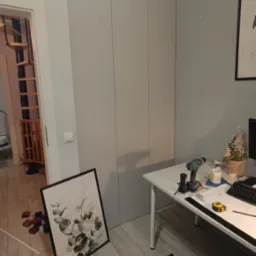 Jasnoszara szafa wnękowa z trzema frontami, widoczna w trakcie montażu z narzędziami na biurku i dekoracją. W tle schody spiralne.