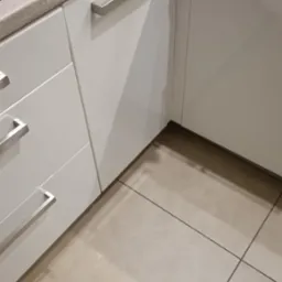 Nowoczesna, biała zabudowa kuchenna z szufladami i szafkami, z długimi, metalowymi uchwytami. Widoczne fragmenty blatu, płyty grzewczej i beżowej podłogi.