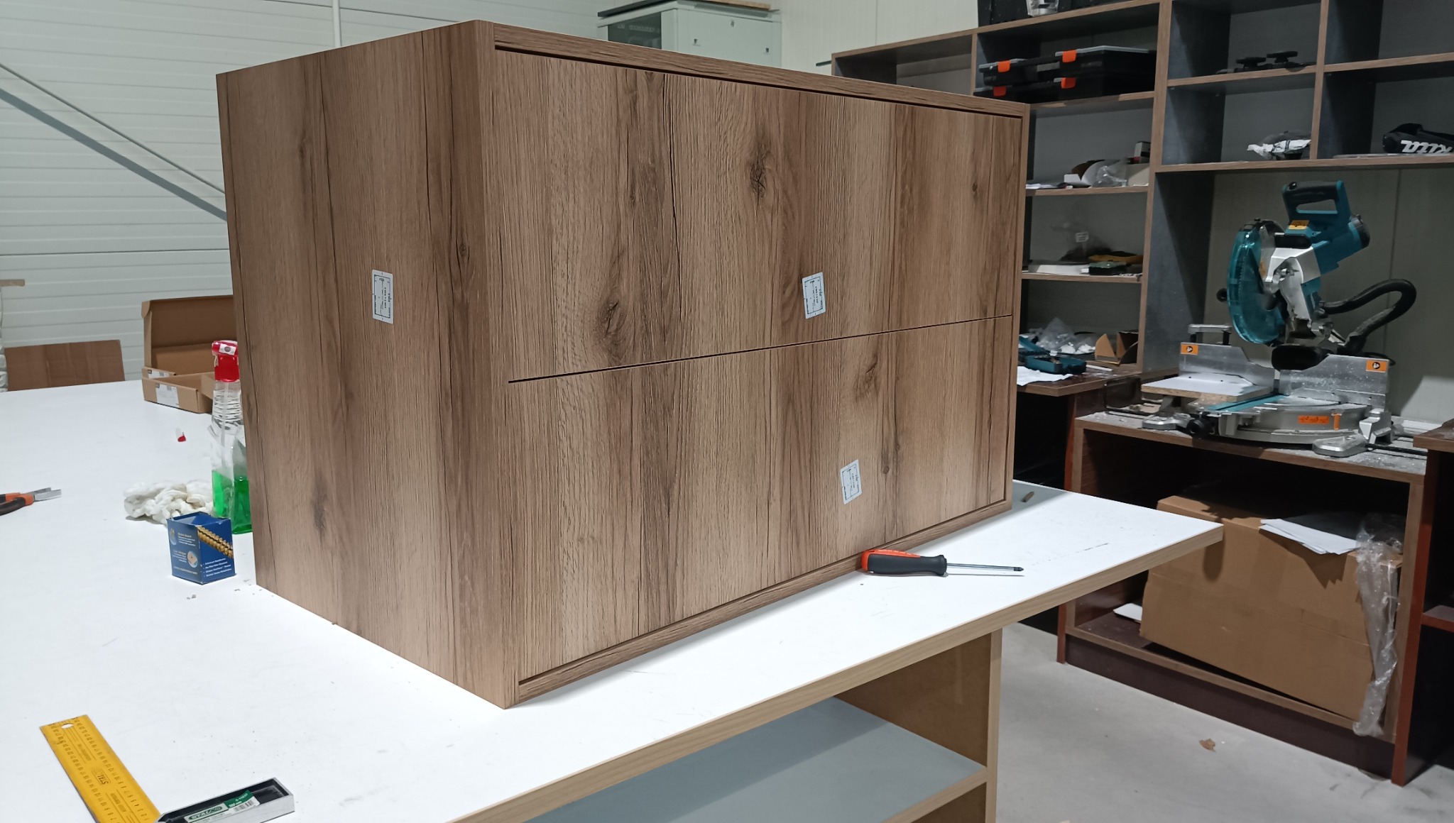 Brązowa szafka z dwoma szufladami o drewnopodobnej fakturze, częściowo zamontowana na warsztatowym stole, z narzędziami w tle. Widoczne etapy montażu.