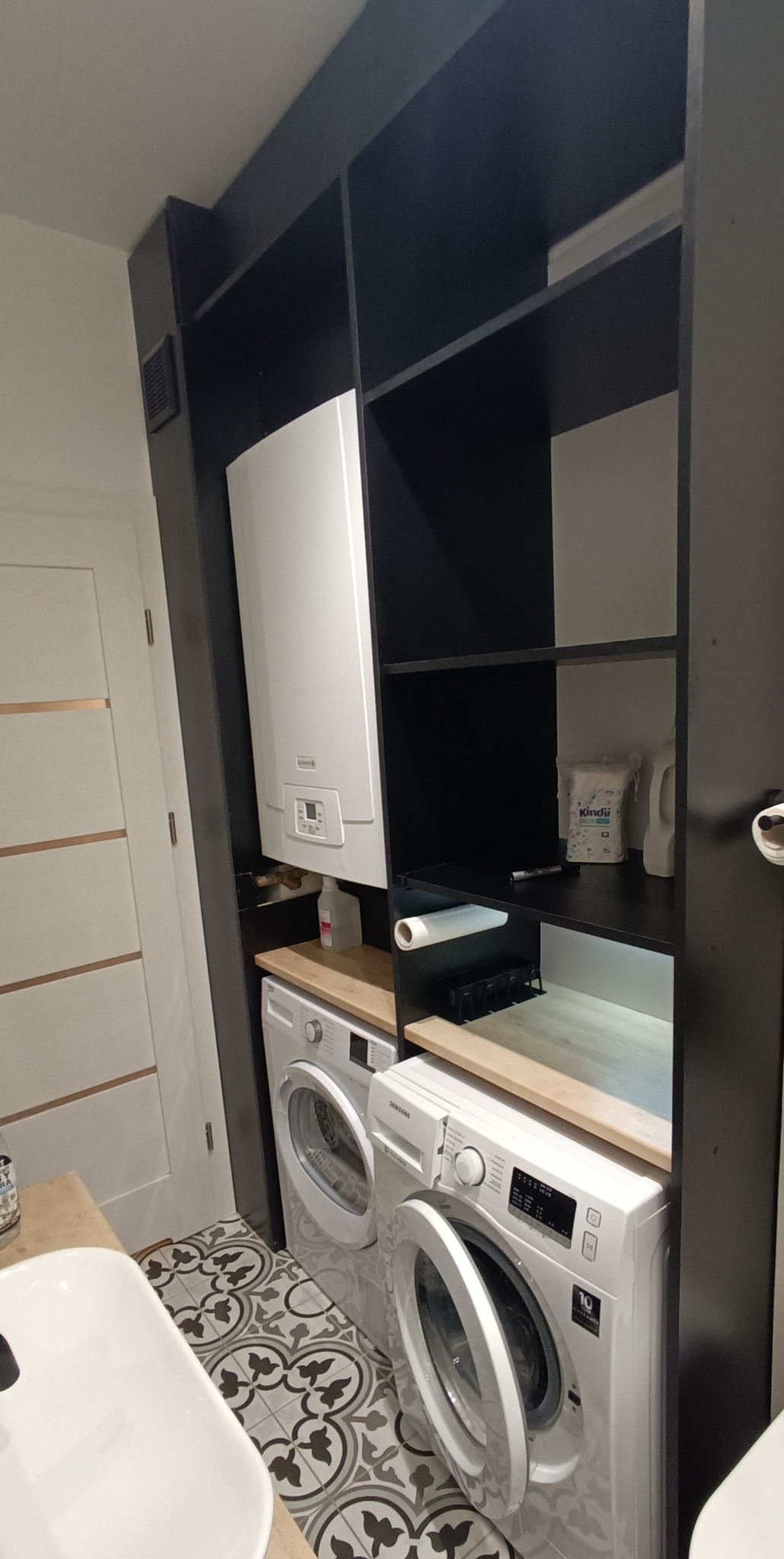 Czarna zabudowa meblowa z półkami, w której umieszczono pralki, bojler i akcesoria łazienkowe. Uwagę zwraca wzorzysta podłoga.