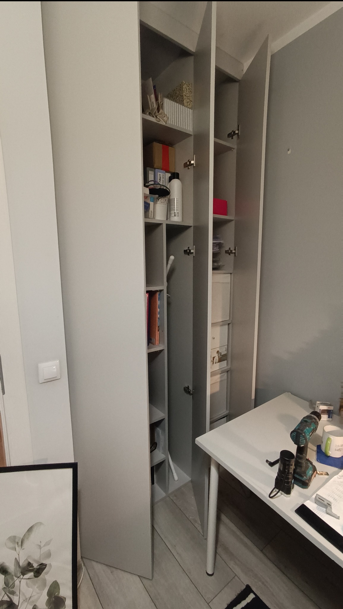 Szara szafa wnękowa z otwartymi drzwiami, widoczne półki z zawartością. Na stole w pobliżu leżą narzędzia, co sugeruje montaż lub prace wykończeniowe.