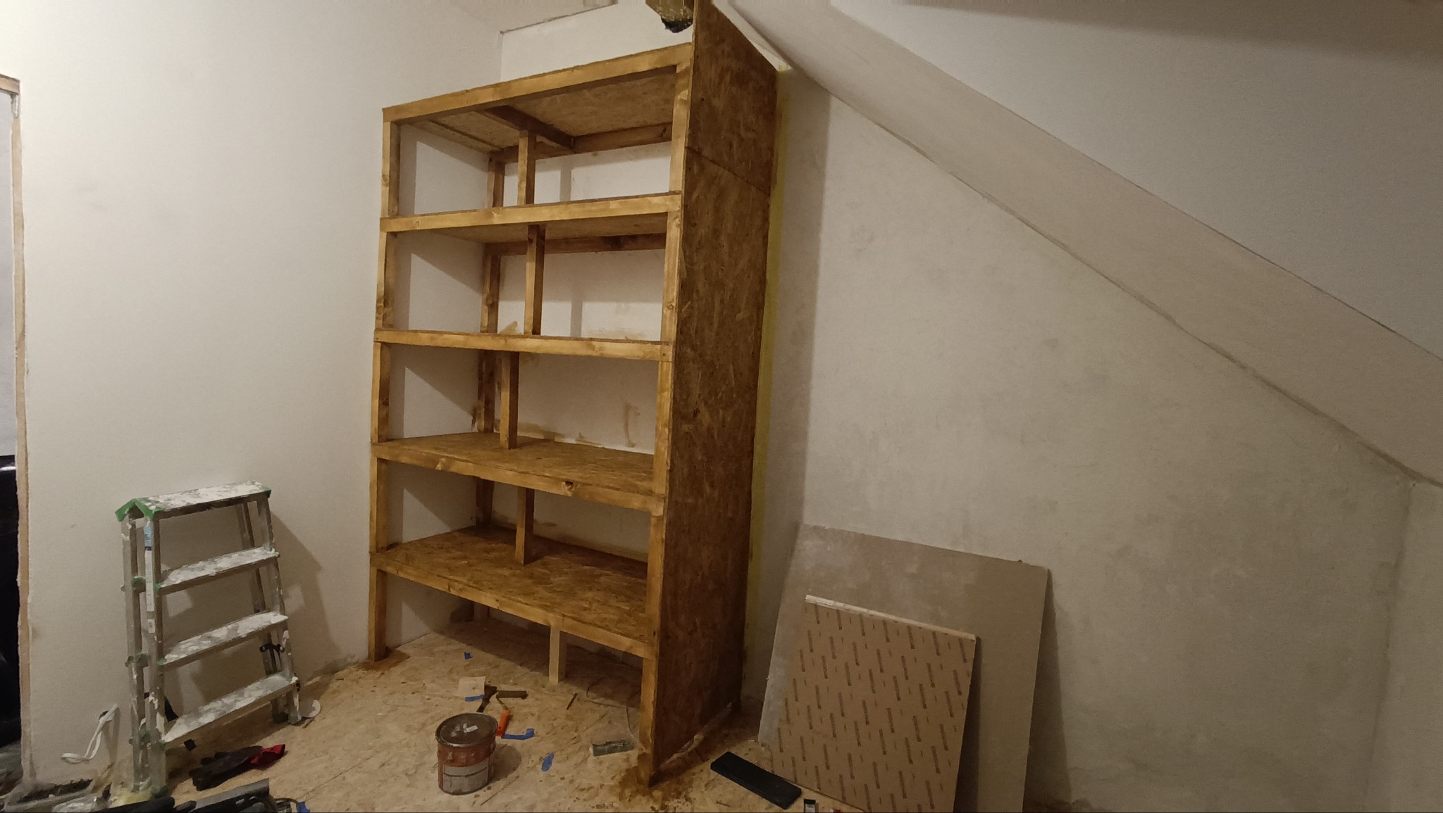 Drewniany regał z surowego drewna, z czterema półkami, w trakcie montażu w pomieszczeniu ze skośnym sufitem. Widoczna drabina i narzędzia.
