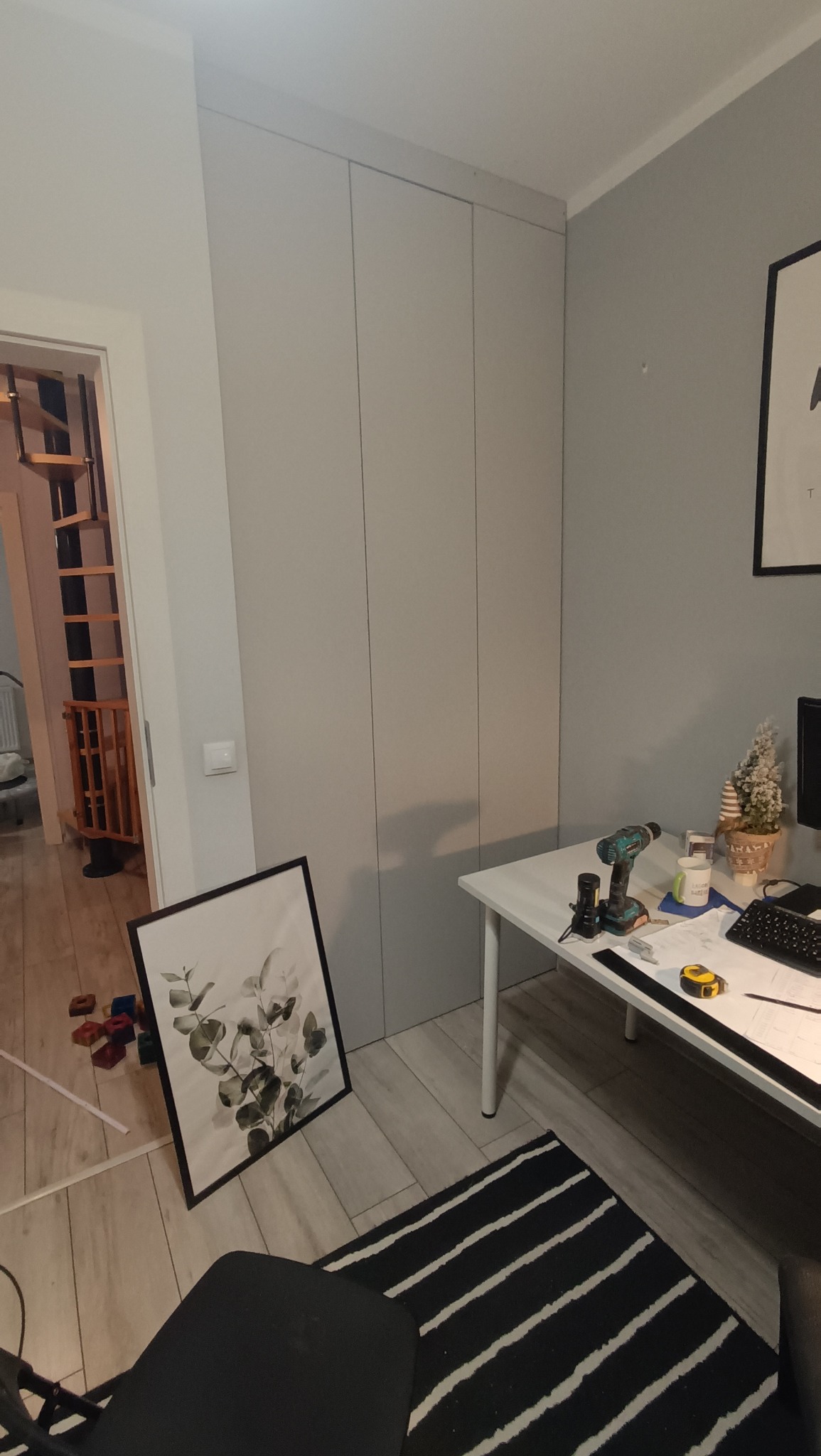 Jasnoszara szafa wnękowa z trzema frontami, widoczna w trakcie montażu z narzędziami na biurku i dekoracją. W tle schody spiralne.