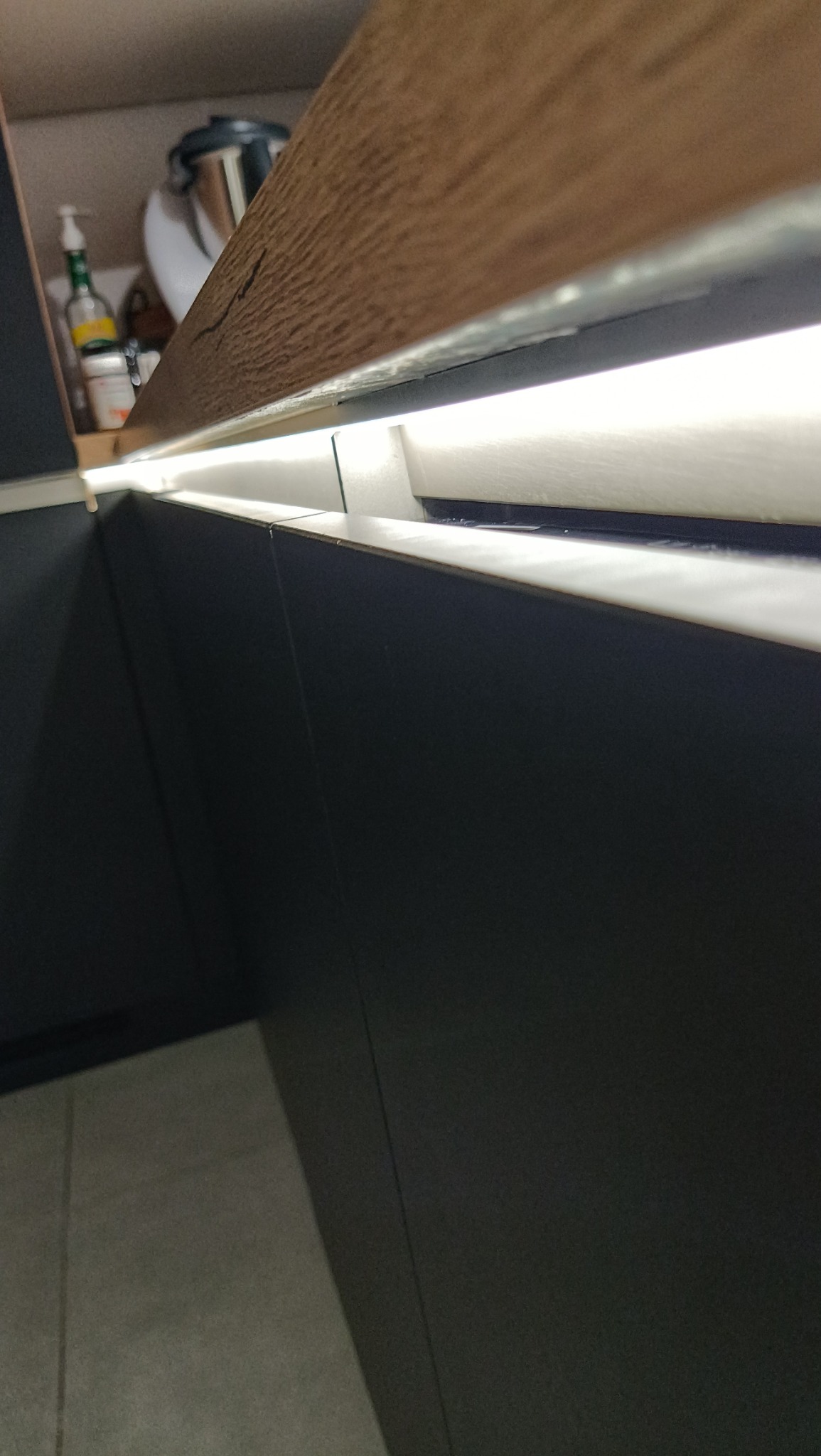 Fragment szafki kuchennej z oświetleniem LED pod blatem z drewnianym wykończeniem. Widoczna część wyposażenia w szafce.