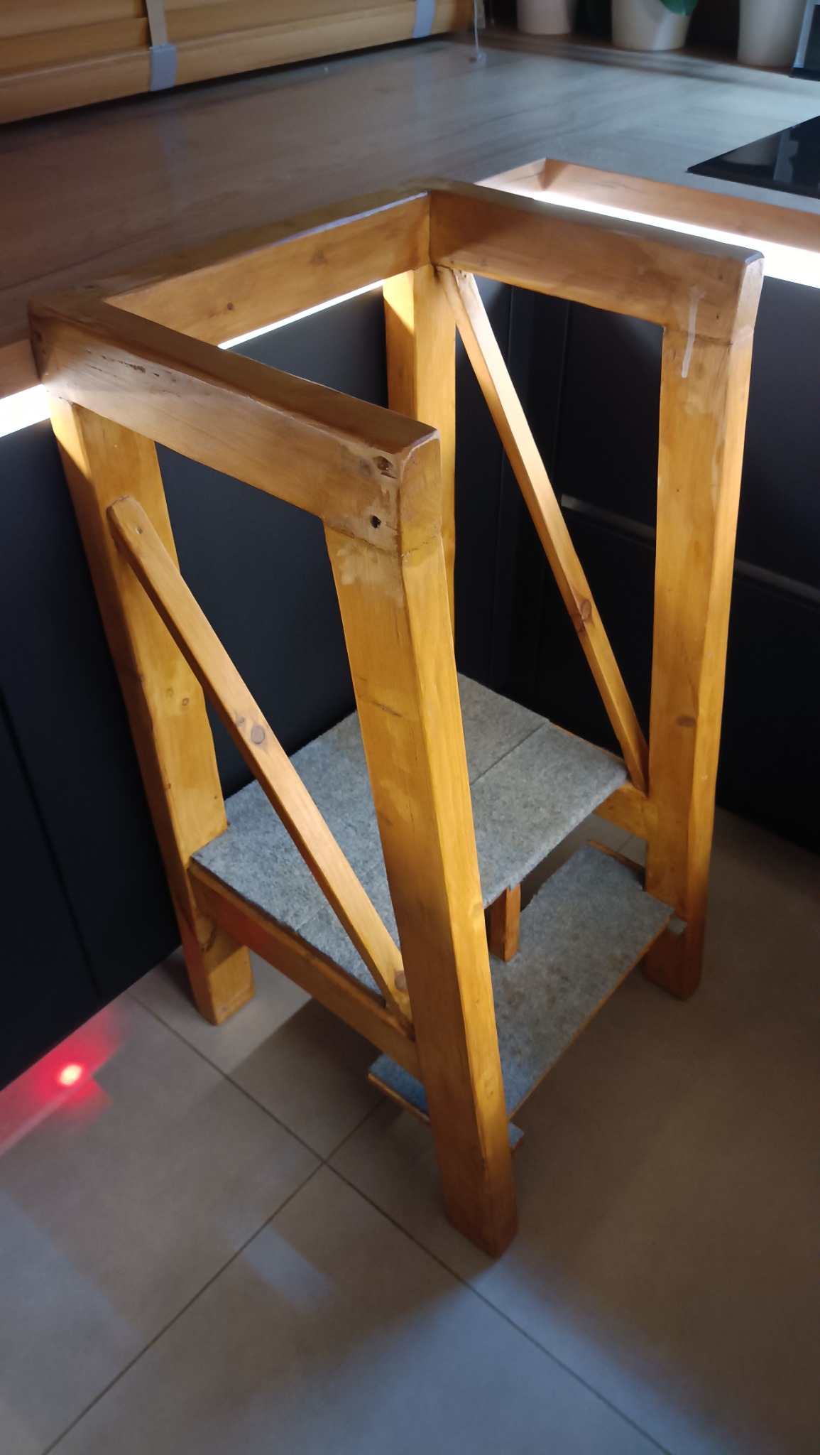 Drewniany podest schodkowy z szarymi stopniami, stojący na płytkach podłogowych, na tle ciemnych szafek kuchennych z oświetleniem podszafkowym.