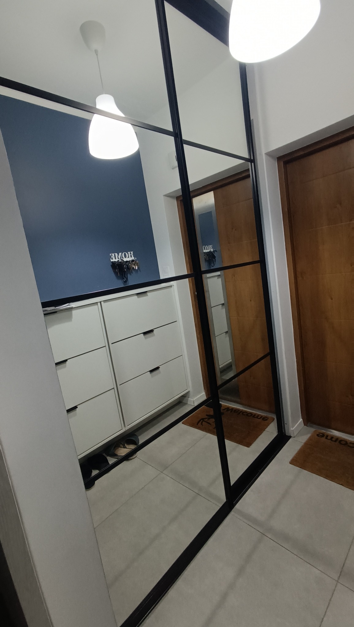 Nowoczesna szafa z lustrem i czarnymi ramami, w tle niebieska ściana z wieszakiem na klucze. Widoczne drzwi i lampy sufitowe. Minimalistyczny design.