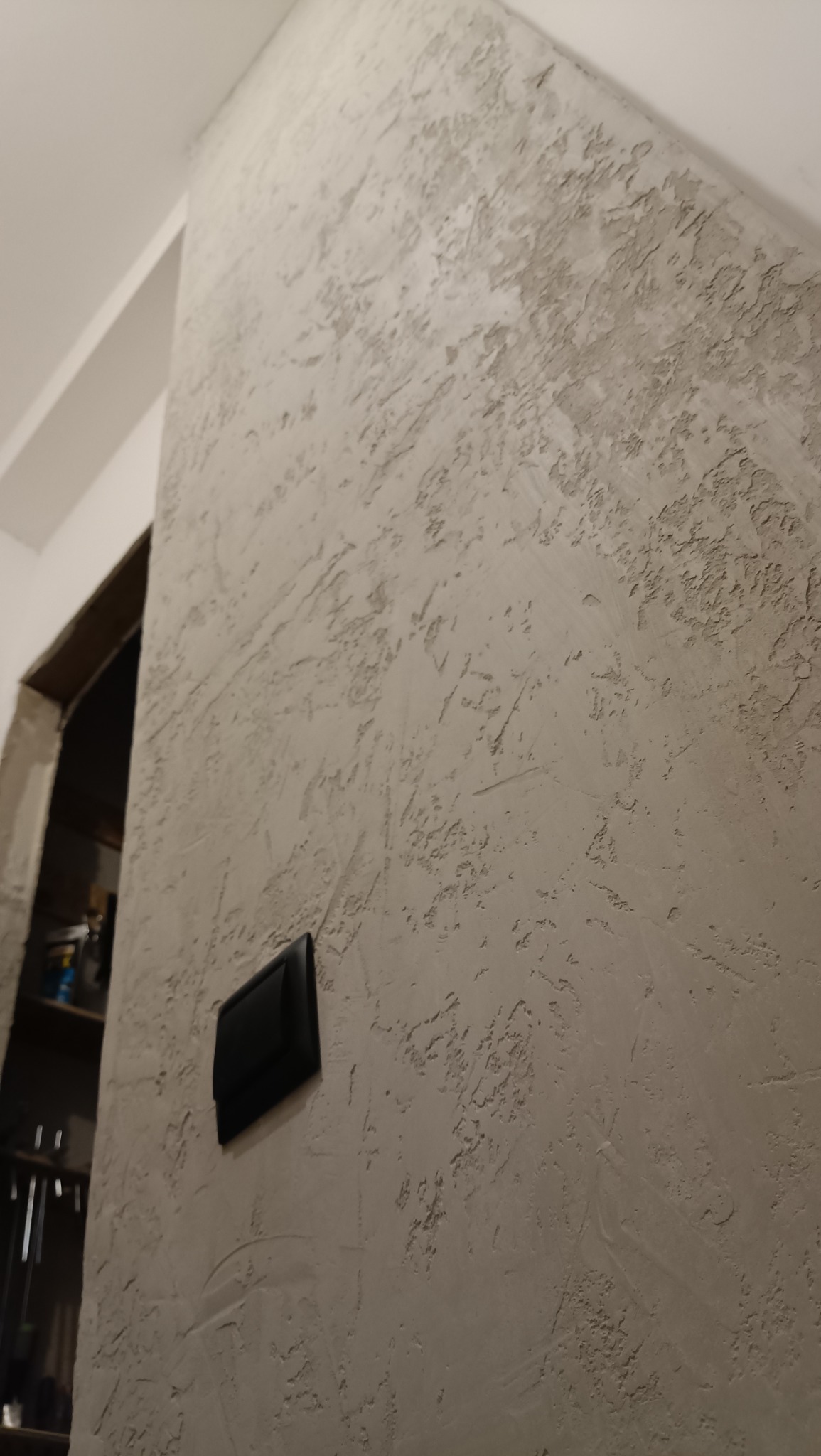 Ściana z szarą, dekoracyjną strukturą tynku, na której zamontowany jest czarny włącznik światła. W tle widoczny fragment półki wnękowej.