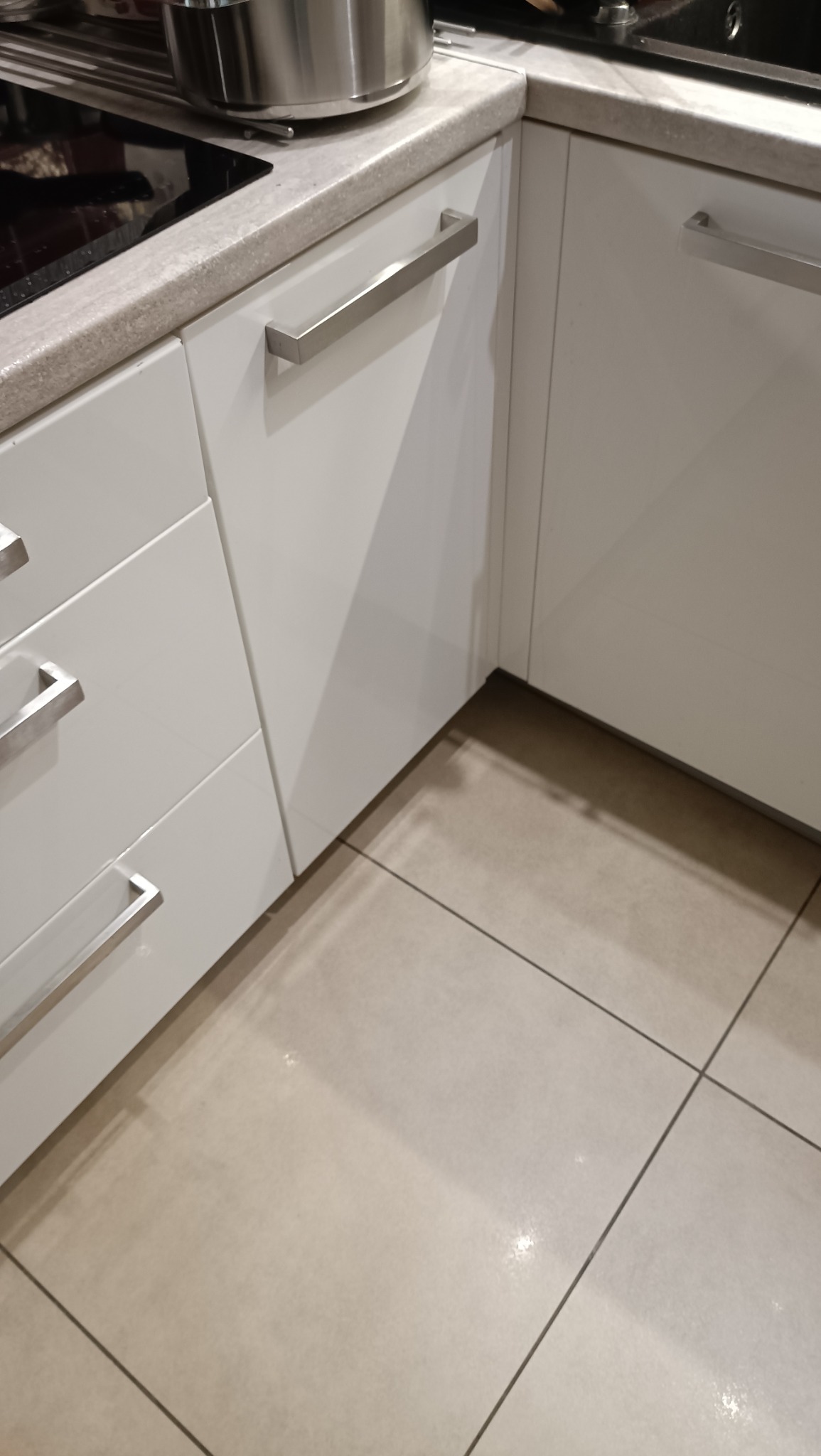 Nowoczesna, biała zabudowa kuchenna z szufladami i szafkami, z długimi, metalowymi uchwytami. Widoczne fragmenty blatu, płyty grzewczej i beżowej podłogi.