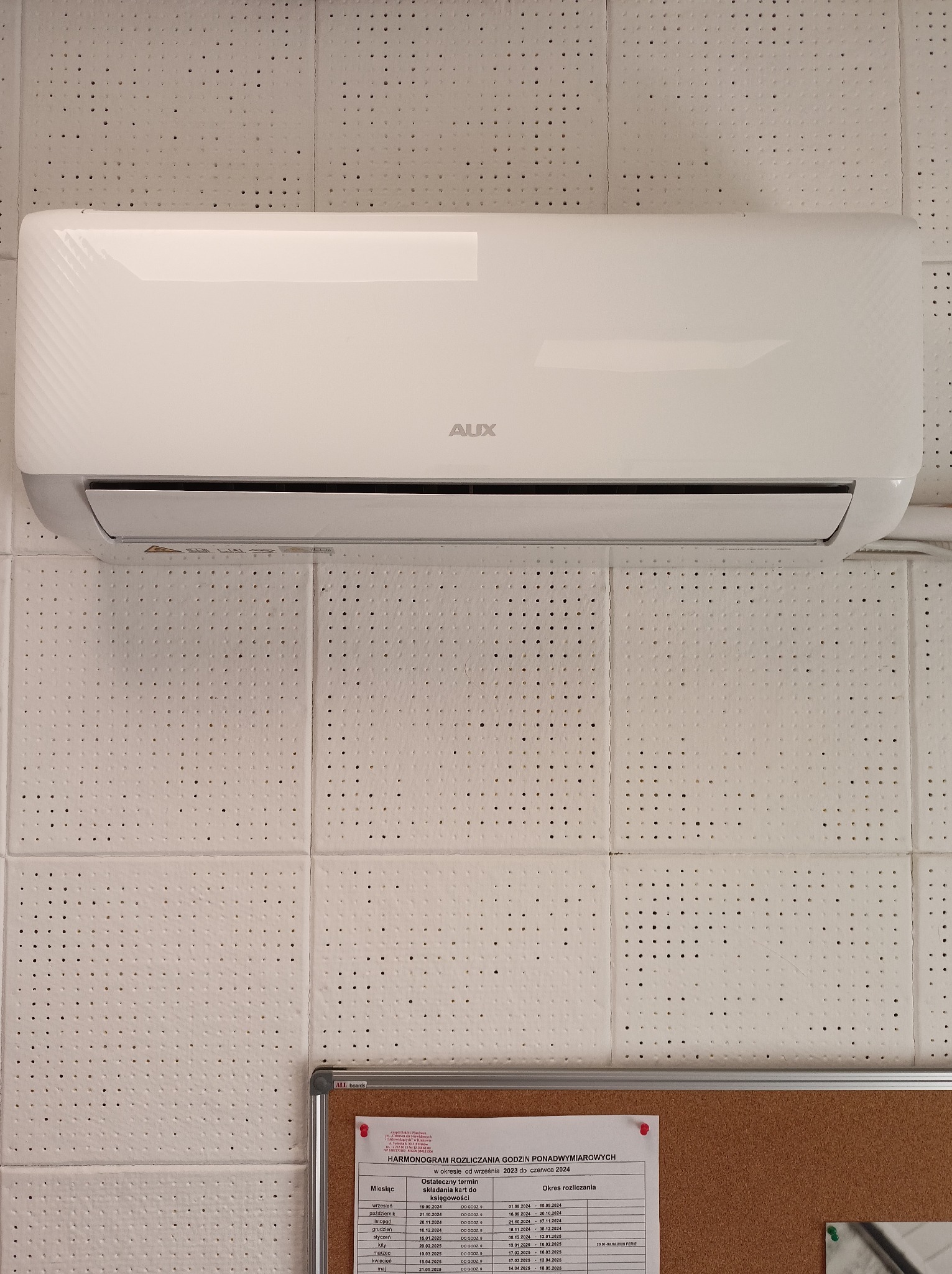 Biały klimatyzator marki AUX zamontowany na ścianie z panelami o perforowanej strukturze. Tablica informacyjna widoczna w dolnej części zdjęcia.