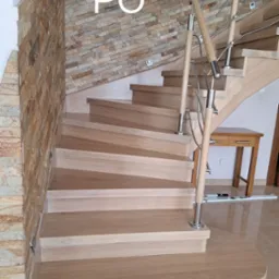 Jasne, drewniane schody po renowacji, z metalową balustradą i kamienną okładziną ściany. Widoczny fragment stolika. Na ścianie napis 'PO'.