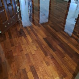 PRO-PODŁOGI - układanie , renowacja - Błyszcząca, świeżo polakierowana podłoga parkietowa z ciemnego drewna, widok z niskiego kąta, z otwartymi drzwiami w tle. Napis 'PO' nałożony na zdjęcie.