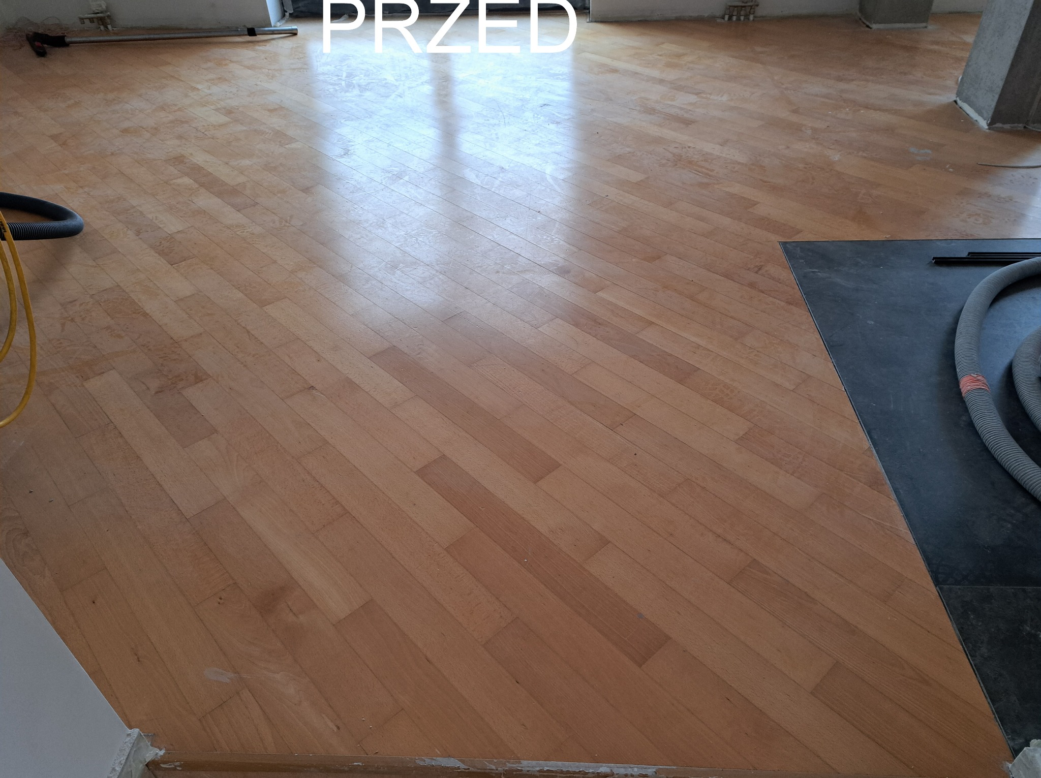 Parkiet bukowy ułożony w jodełkę, widoczny w trakcie prac przygotowawczych z napisem 'PRZED'. Fragment odkurzacza przemysłowego w rogu.