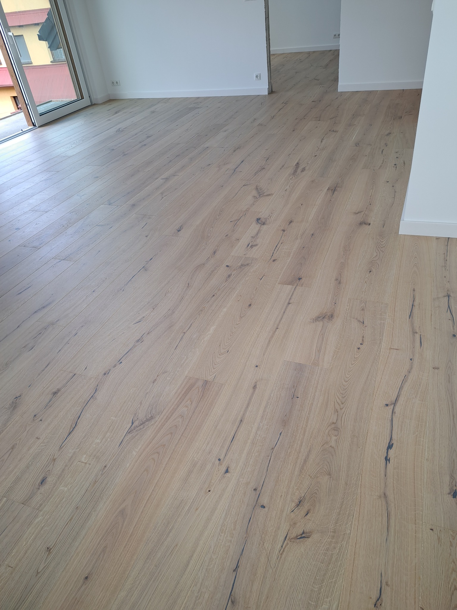 Jasne panele podłogowe z wyraźnym rysunkiem drewna, ułożone w nowym, jasnym pomieszczeniu z widokiem na balkon i inne pokoje.