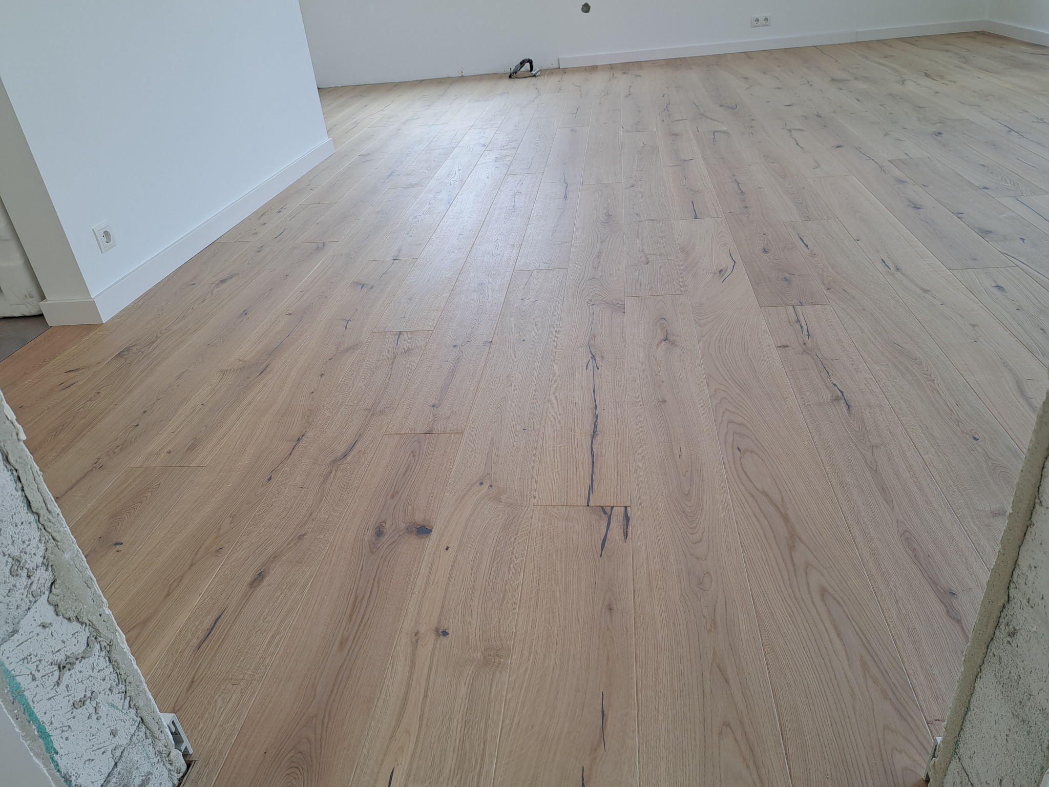 Jasny, dębowy parkiet ułożony w przestronnym wnętrzu. Widoczne subtelne słoje drewna i minimalistyczne listwy przypodłogowe. Fragment surowej ściany w tle.