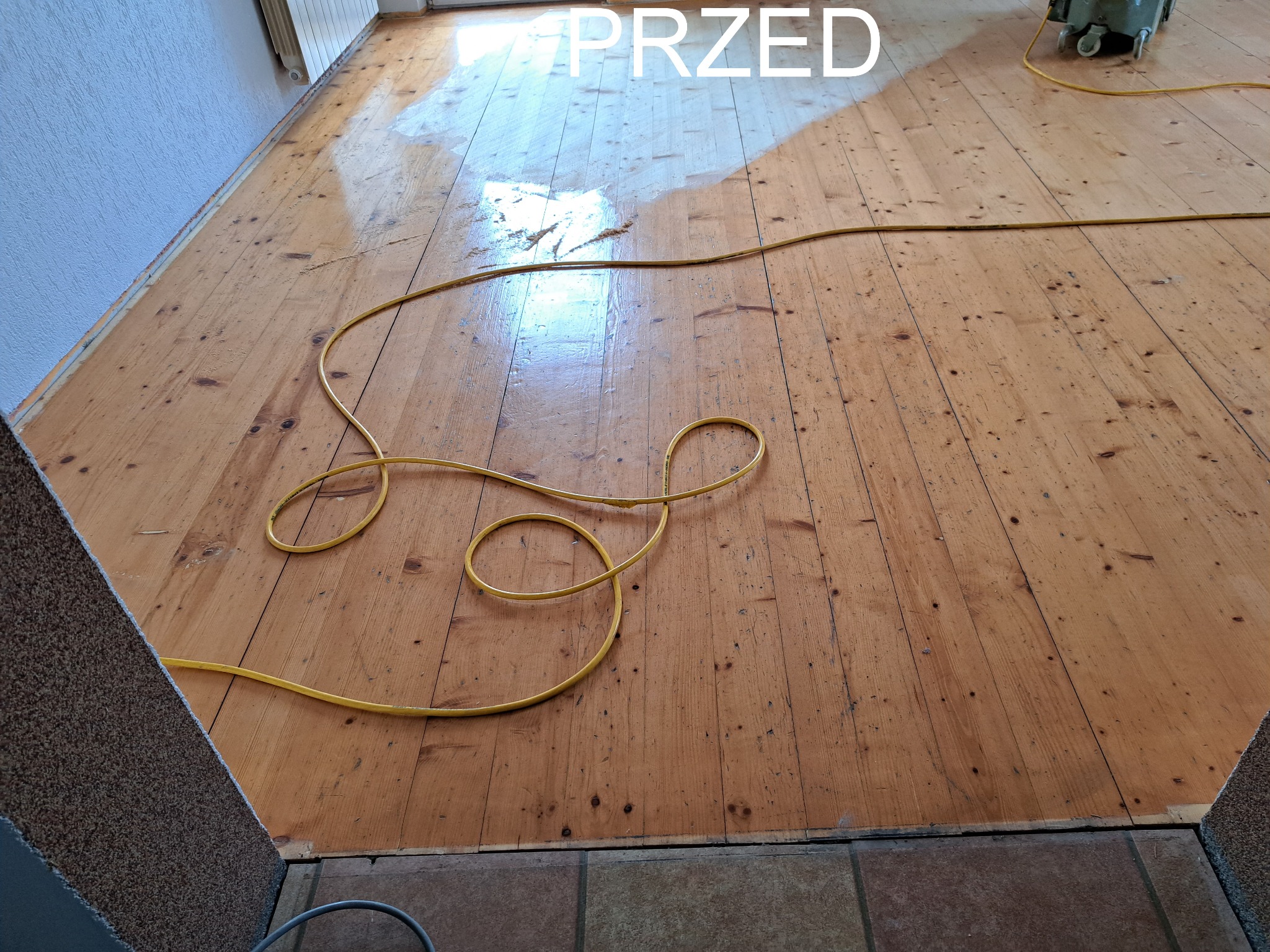 Parkiet przed cyklinowaniem: widoczne ślady użytkowania, rysy i zabrudzenia. Żółty kabel od maszyny leży na podłodze. Część podłogi już została wyczyszczona.