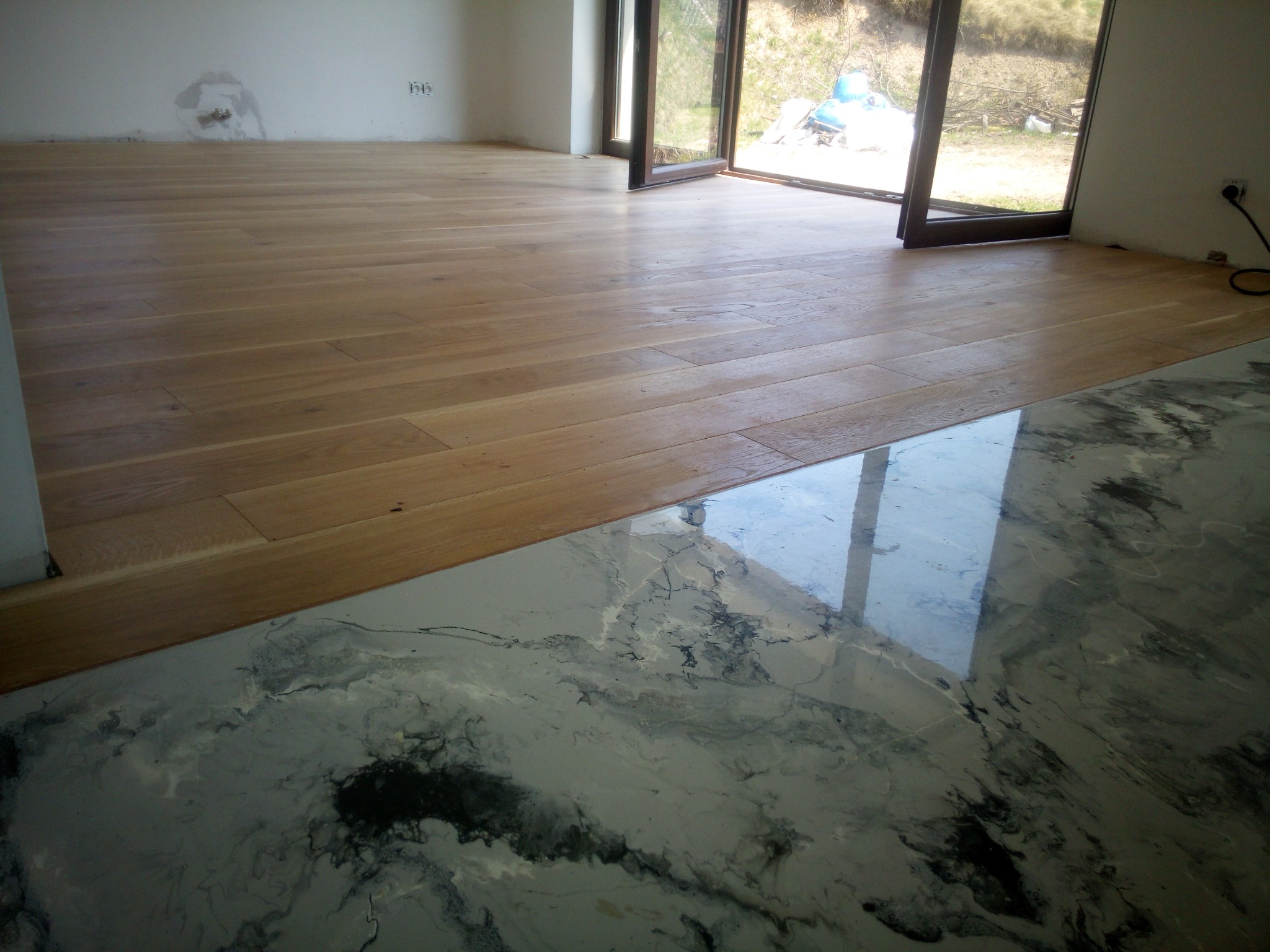 Fragment wnętrza: parkiet z jasnego drewna ułożony obok posadzki z żywicy epoksydowej o wzorze marmuru. Widoczne okno balkonowe i fragment ściany z gniazdkami.