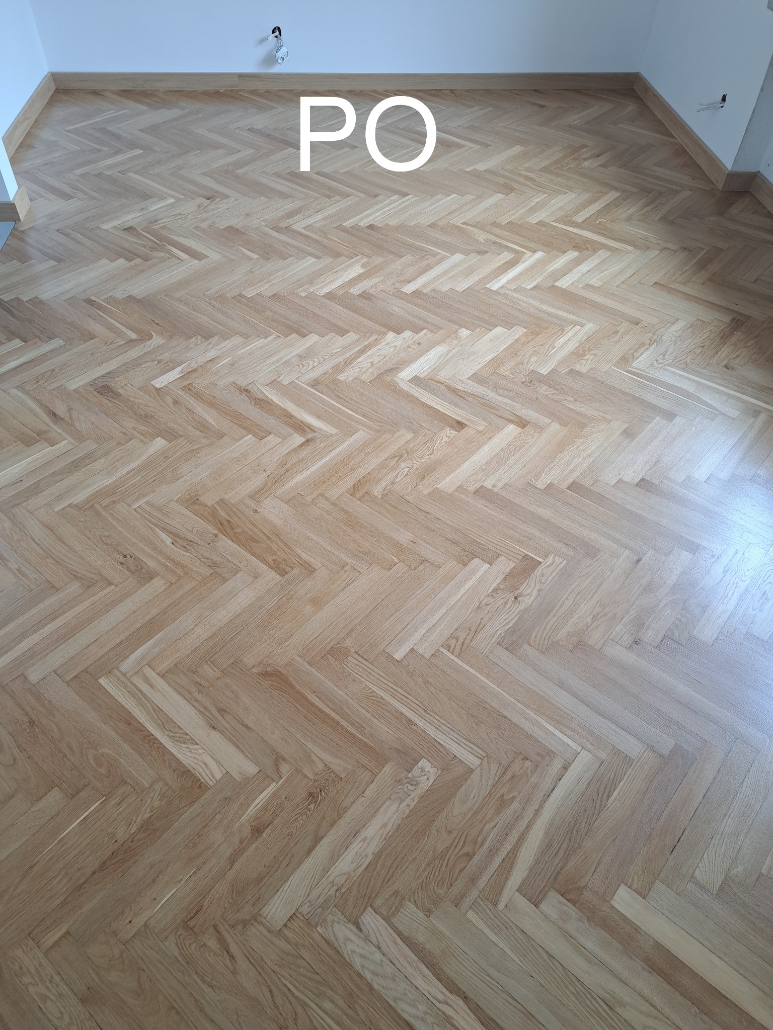 Jasny parkiet ułożony w jodełkę po renowacji, widok z góry. Na podłodze widoczny napis 'PO', co sugeruje etap po wykonanej usłudze.