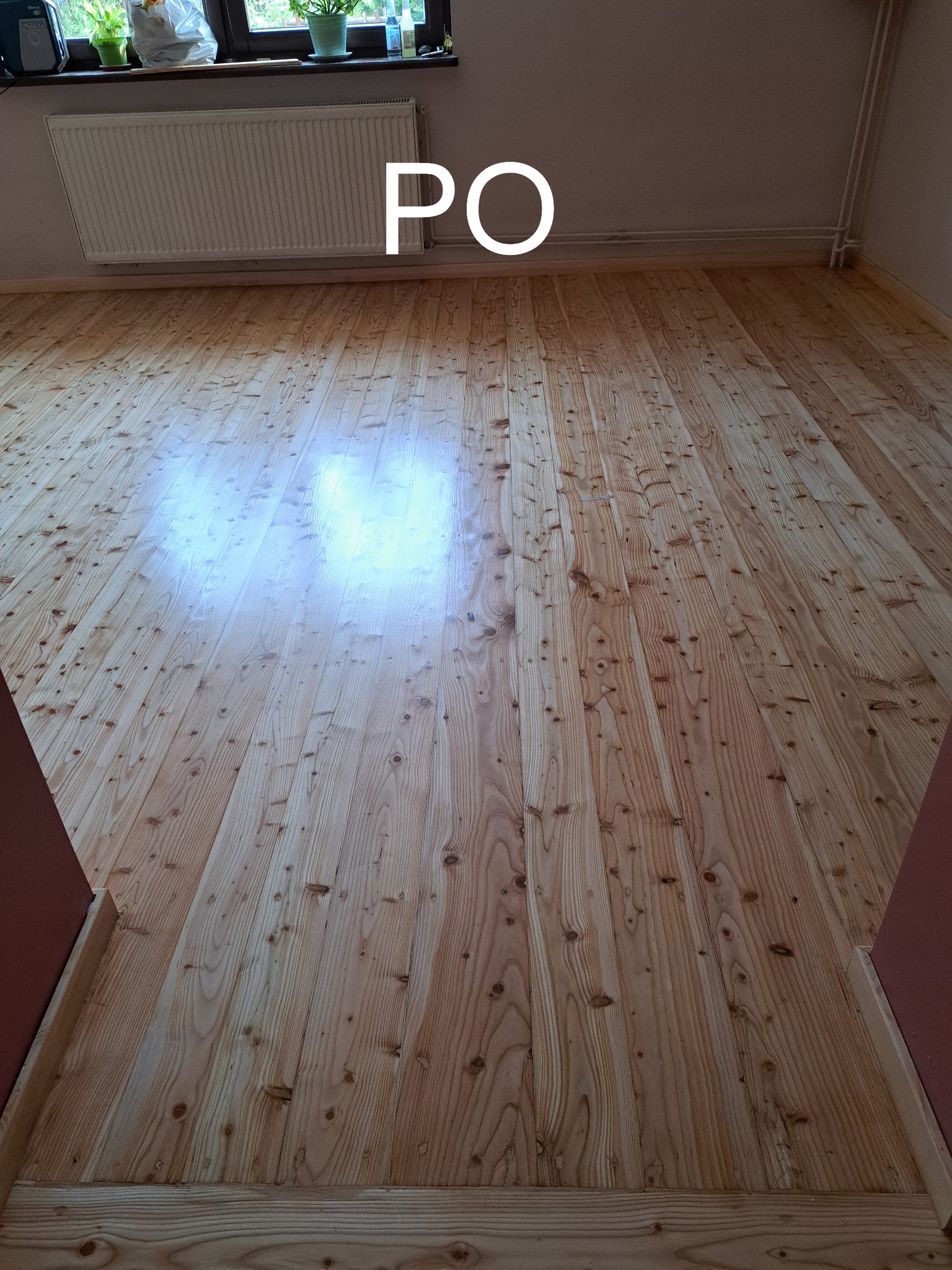 Jasne, odnowione deski podłogowe z widocznym rysunkiem drewna po cyklinowaniu, z białym napisem 'PO'. Na ścianie grzejnik.
